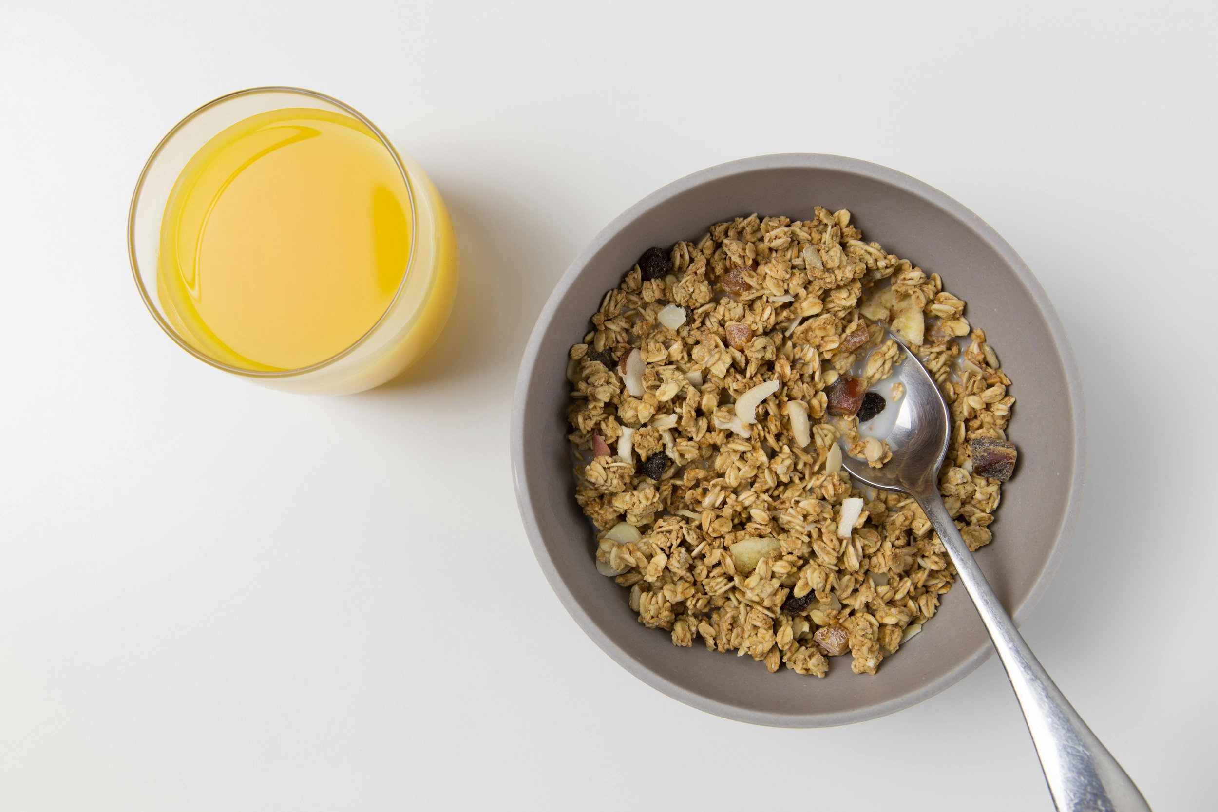 Top-down view of a bowl of granola with dried fruit and a spoon, placed next to a glass of orange juice on a white surface.