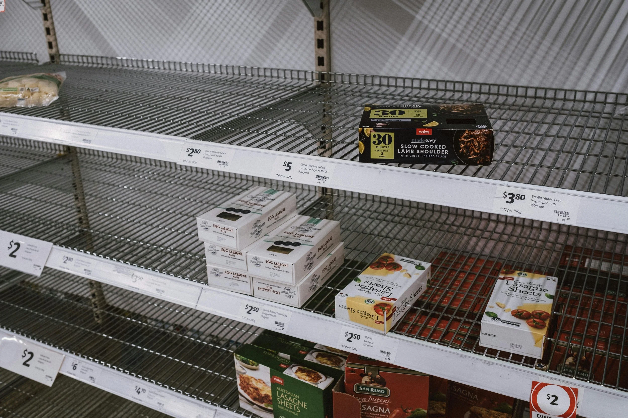 Nearly empty supermarket shelves with a few boxed food items remaining, showing product prices and gaps where stock is missing.