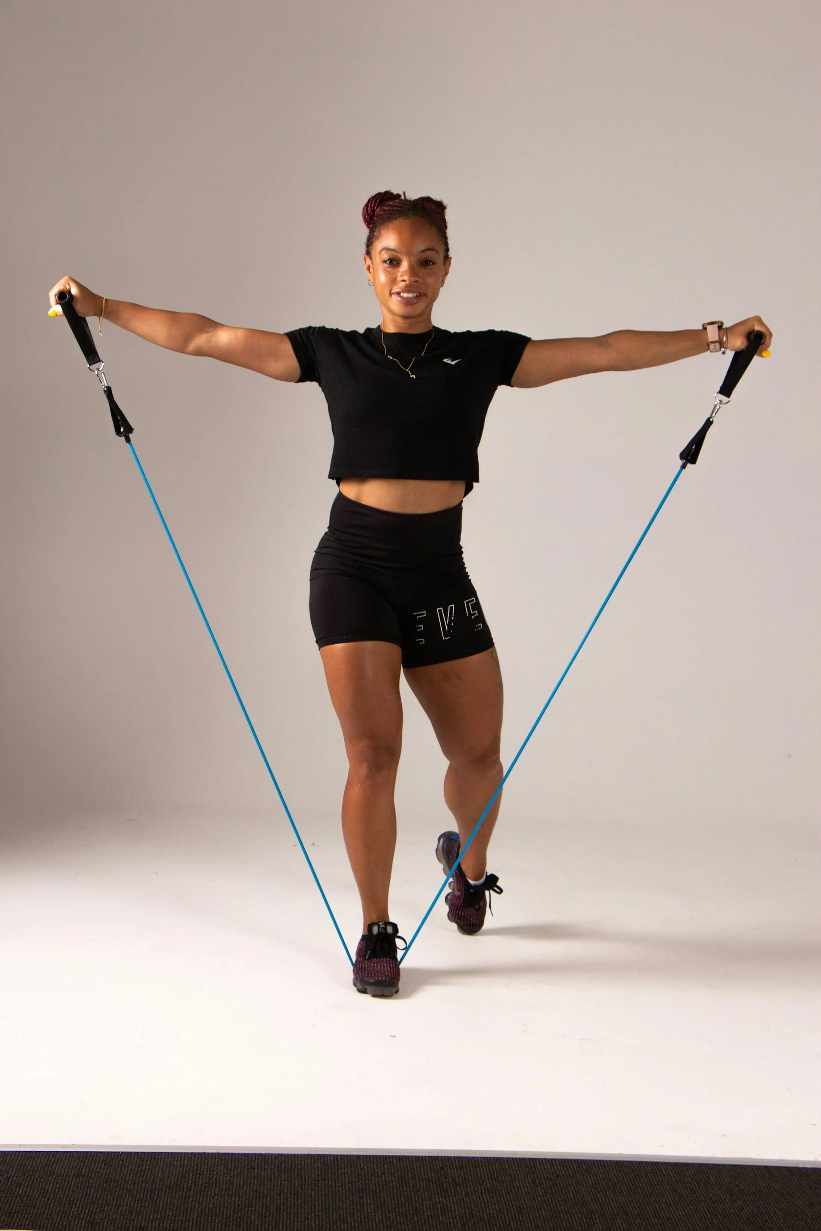 A woman is exercising with a resistance band, standing on one leg, with arms outstretched holding the band, indoors against a plain wall.
