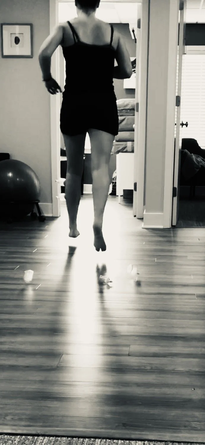 A woman in a short black dress is jumping in the air inside a house, viewed from behind, with a reflection visible on the wooden floor.