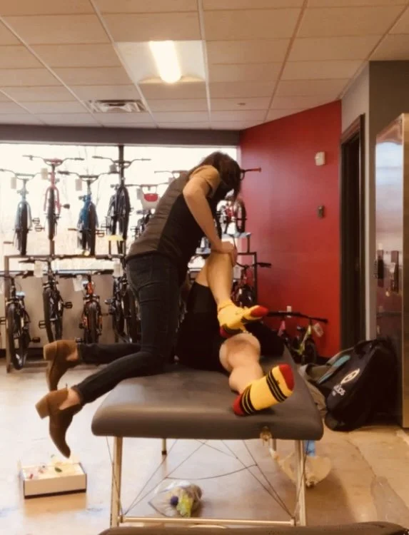 A person performs a mock chiropractic adjustment on a man lying face up on a treatment table in a bicycle shop, with bicycles displayed on the wall behind them.