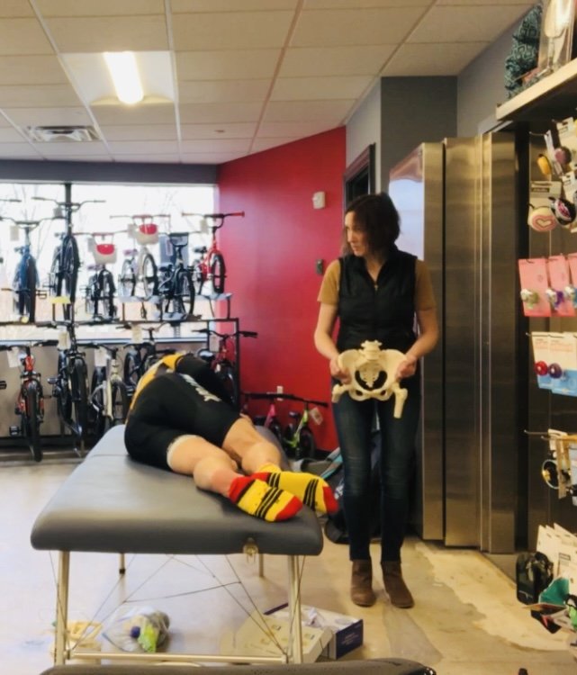 A person lying face down on a massage table with their head turned to the side, wearing a black shirt, yellow socks with red and black stripes, and black shorts. A woman standing next to the table holds a plush skull toy in a pet carrier. The scene is inside a bike shop with multiple bicycles displayed in the background, a red accent wall, and shelves with various items.