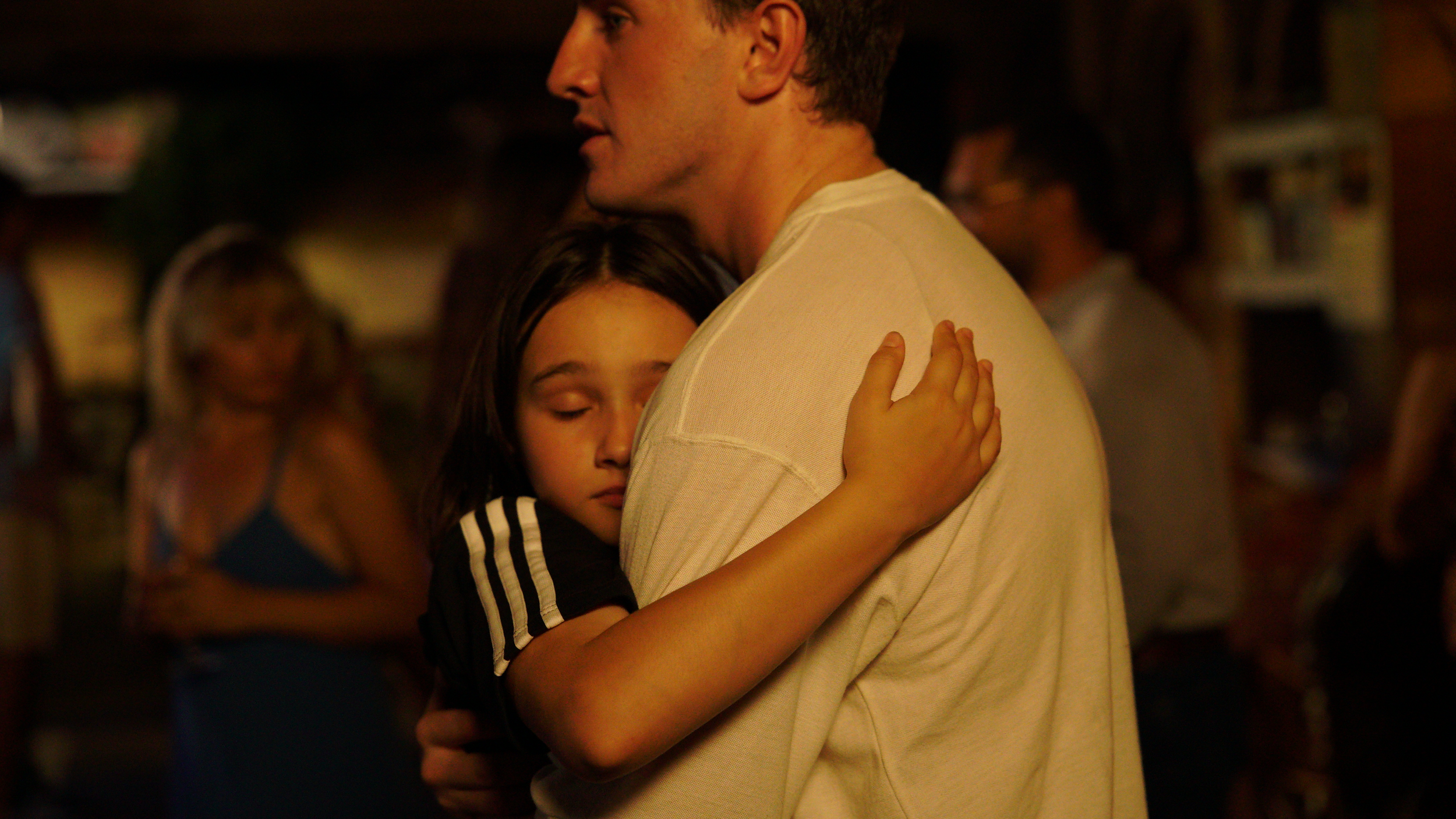 A young girl with closed eyes hugging a man, possibly her father, in an embrace in a dimly lit room with blurred people in the background.