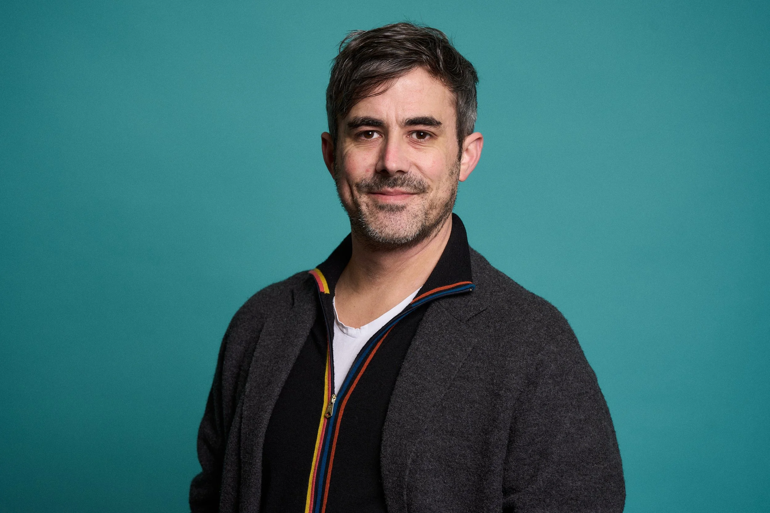 A man with short dark hair and a beard, wearing a black jacket with colorful zipper stripes over a white shirt, smiling softly against a teal background.