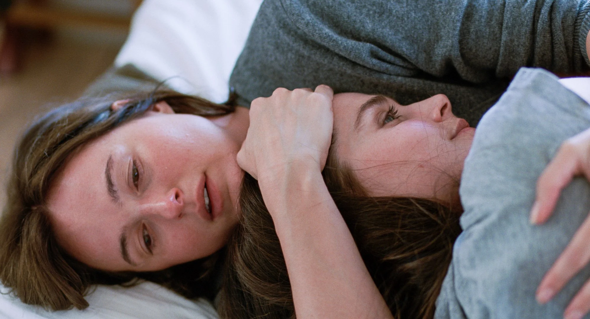 Two women lying on the floor, one with brown hair and the other with light brown hair, appear distressed or upset, with one holding the other's face gently.