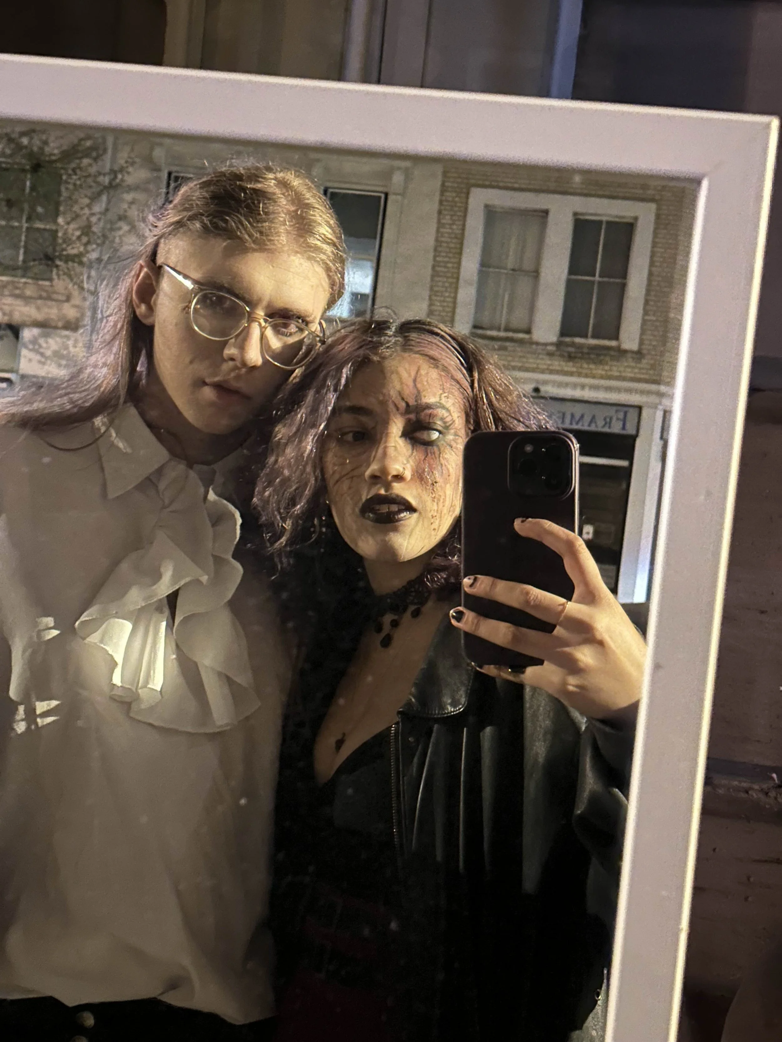 A couple taking a selfie in front of a mirror outdoors at night. One woman has glasses and a white blouse with ruffles, the guy has makeup and a leather jacket.