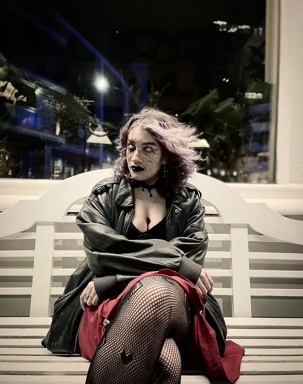 A woman with pale makeup, dark lipstick, and purple hair sitting on a white bench at night. She is wearing a leather jacket, fishnet stockings, and a red skirt, with dark eye makeup and accessories. The background features a window with plants and reflections of lights.