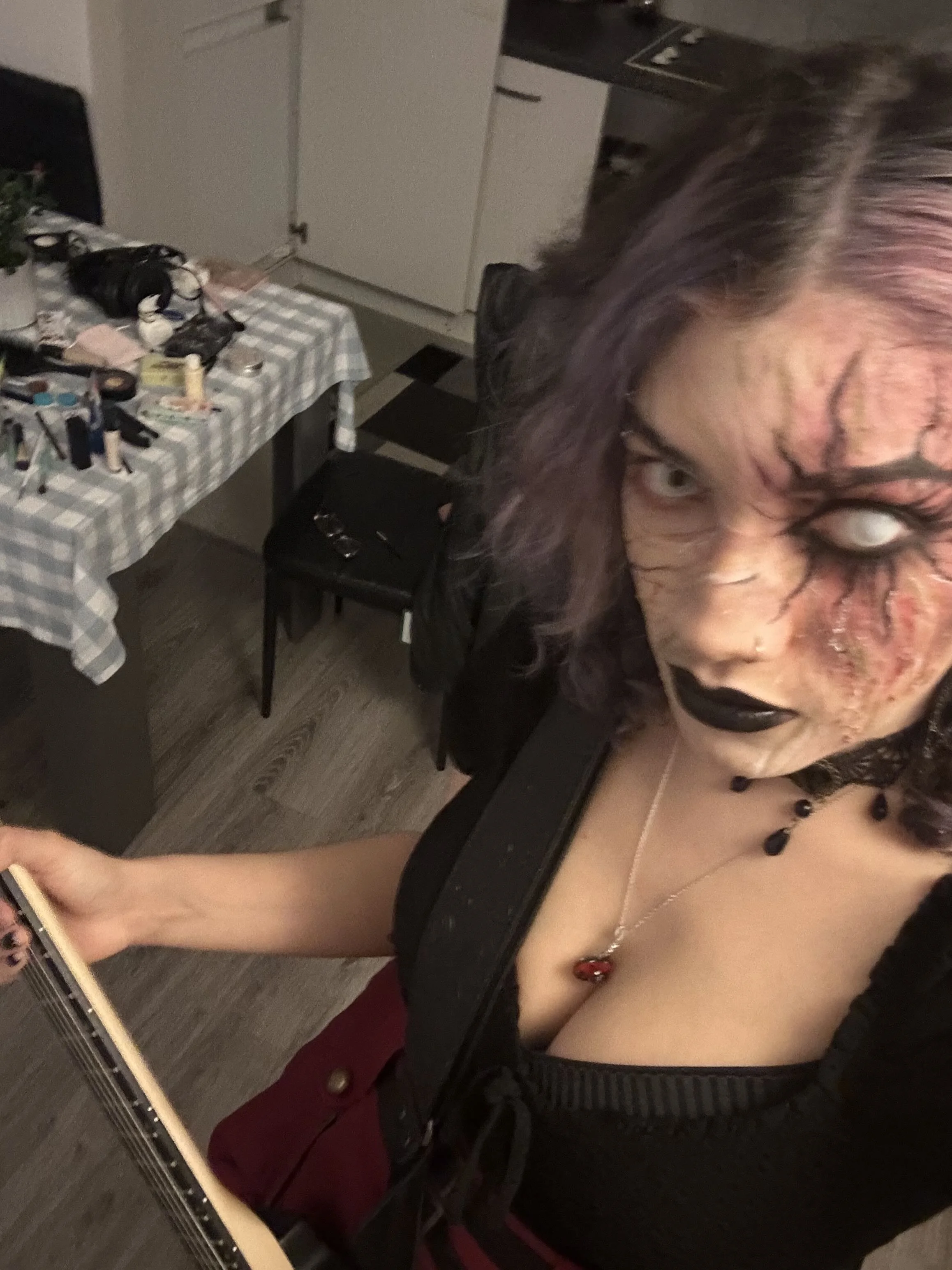 Person wearing gothic makeup and black lipstick, holding a guitar in a room with a table cluttered with makeup and personal items.