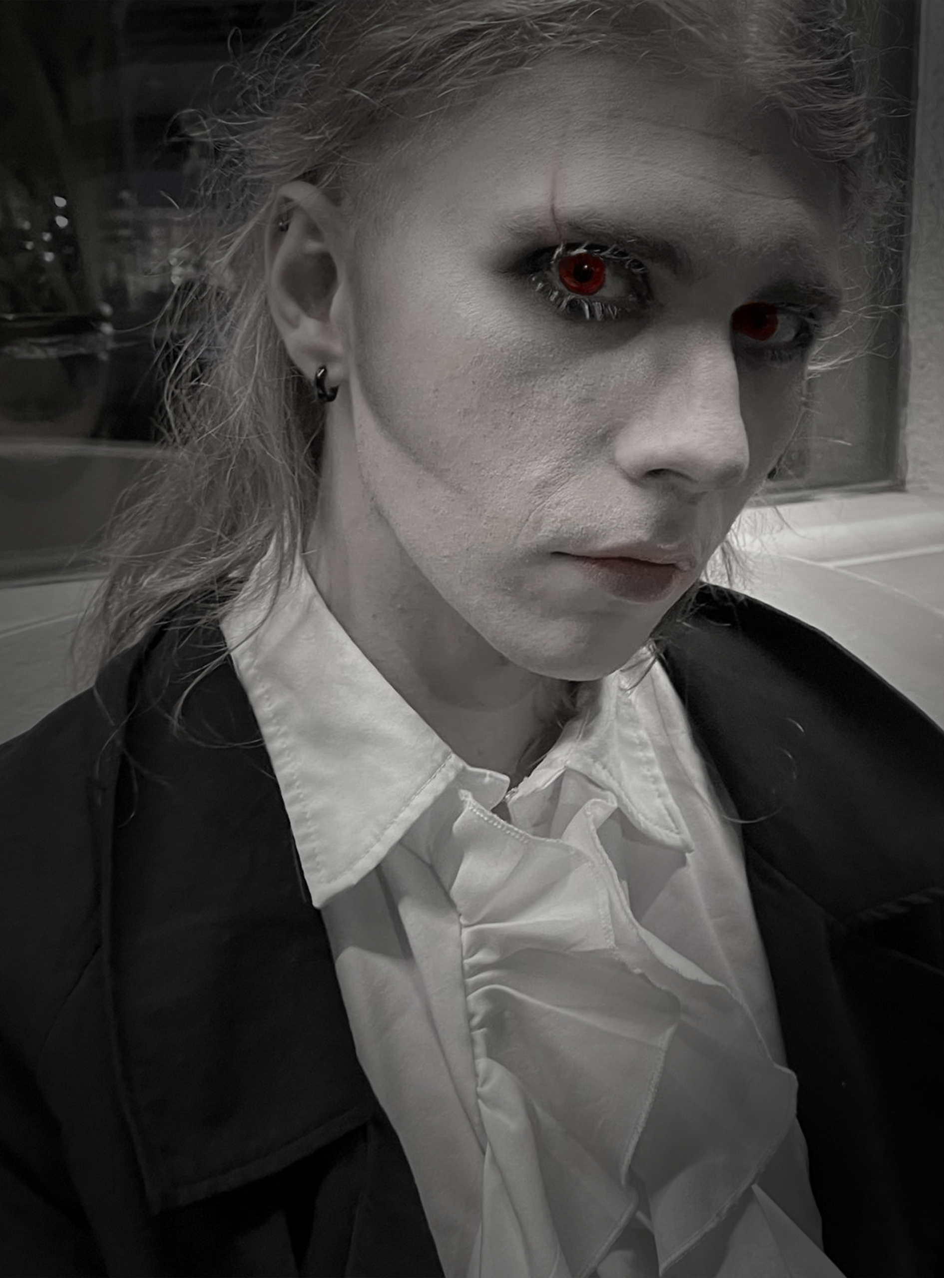 Black and white photo of a person with light-colored hair, pale skin, dark eye makeup, and red-colored eyes, wearing a white ruffled shirt and a dark jacket.