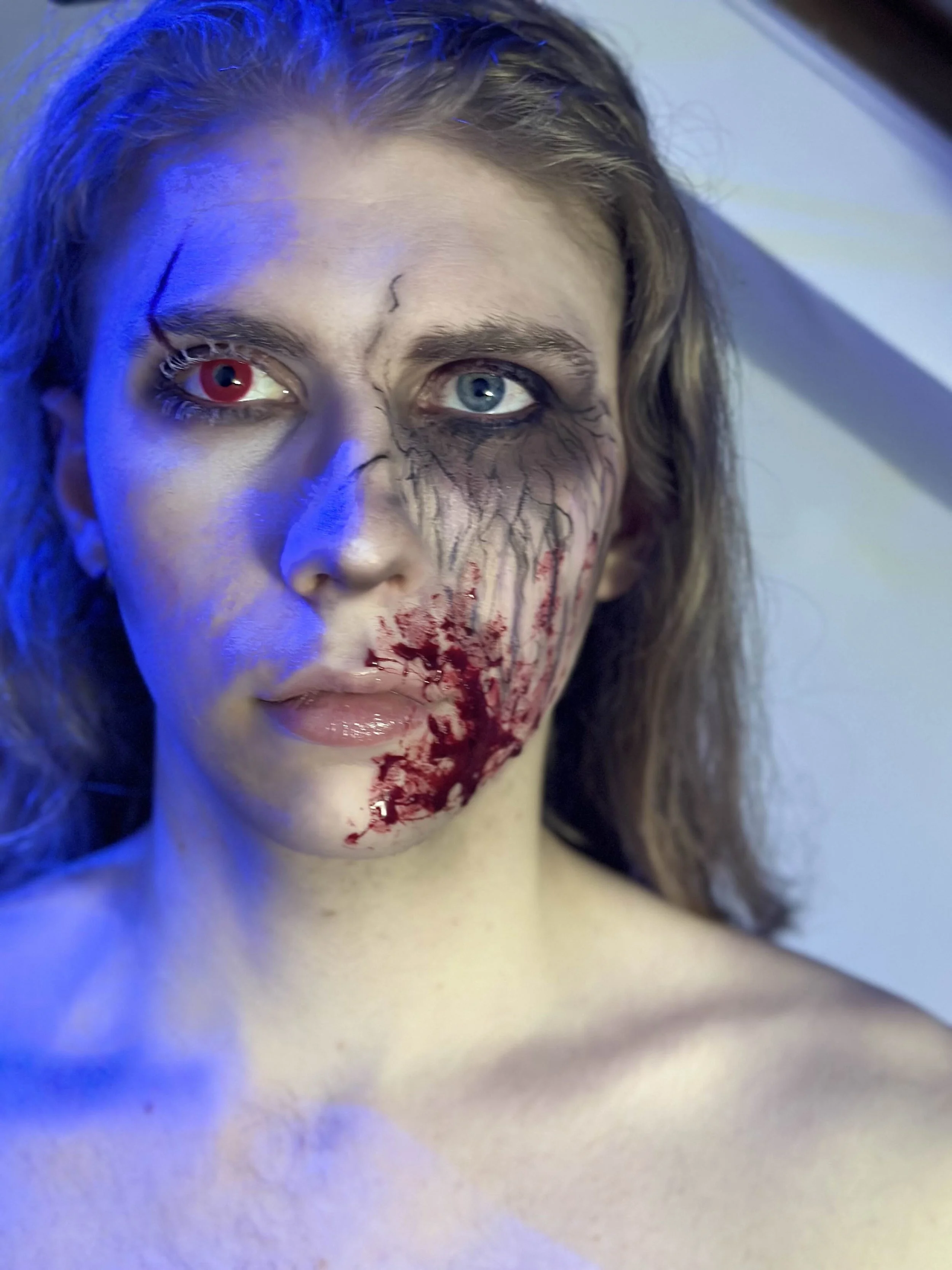 A person with half decayed makeup face, with one eye red and the other blue, and blood and scars around the mouth.