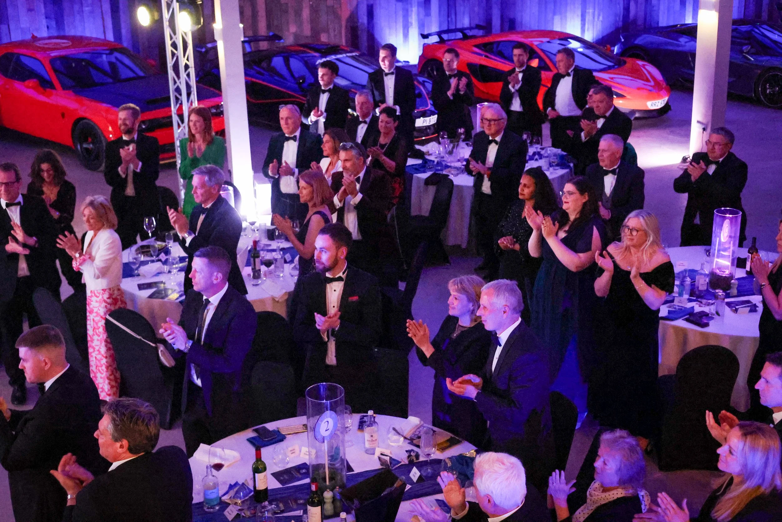 A banquet hall with people dressed in formal attire, many clapping, and several luxury sports cars on display in the background.