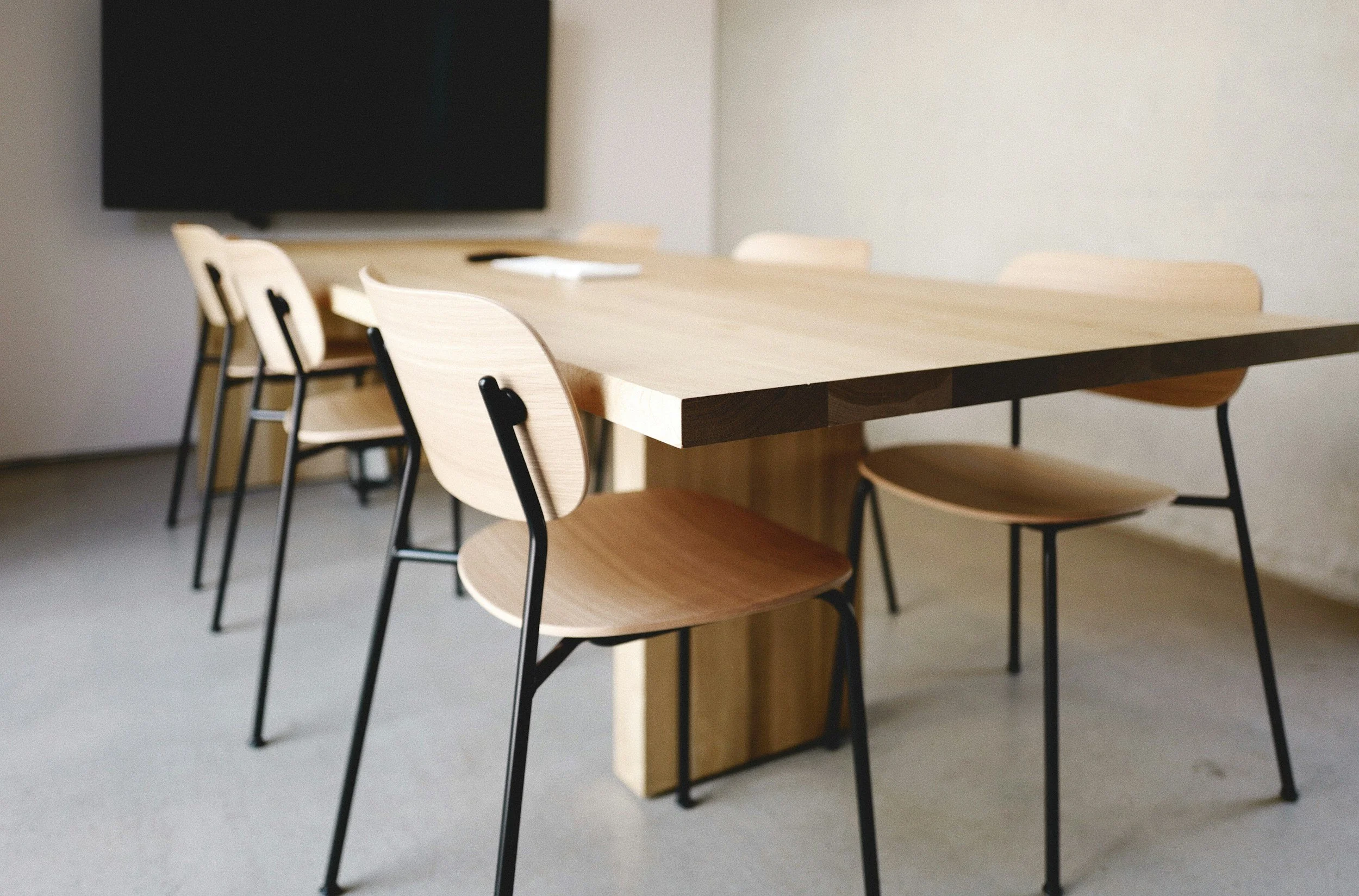 Modern conference room with a large wooden table and six matching wooden chairs with black metal legs, and a black rectangular wall-mounted panel or screen in the background.