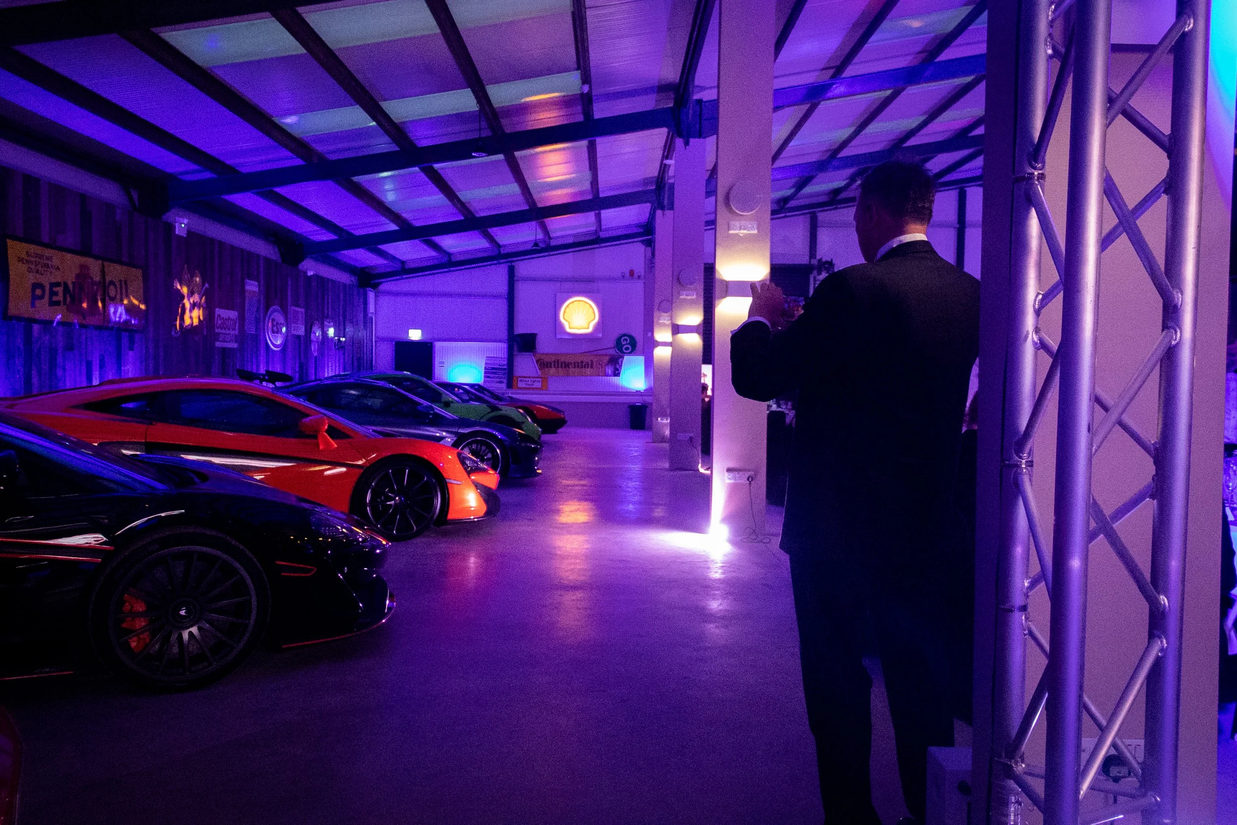 Indoor showroom with luxury sports cars under purple and blue lighting, a man in a suit taking a photo, Shell logo illuminated on the wall, vintage Pennzoil and Castrol signs.