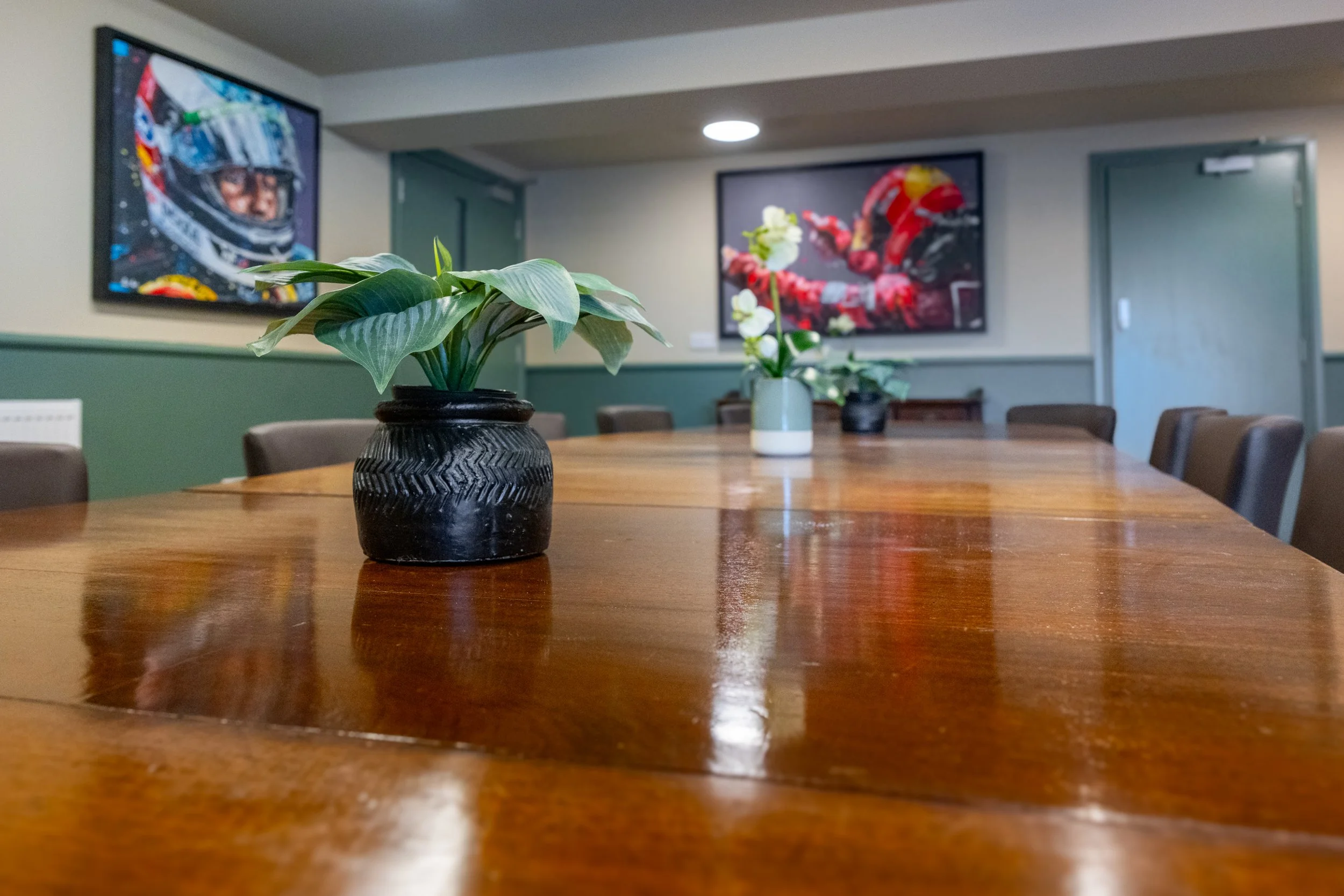 A wooden conference table with three potted plants on it, positioned against a wall with two framed pictures of race car drivers.