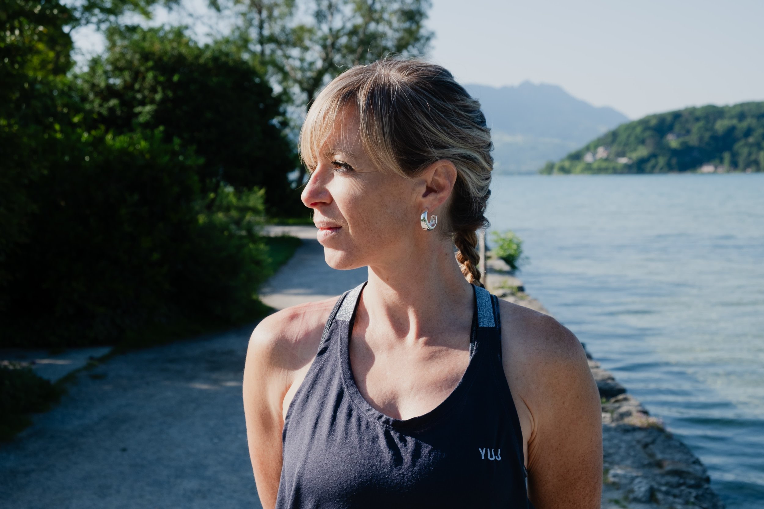 Femme regardant vers la gauche en tenue de sport au bord d'un lac, avec des montagnes en arrière-plan, par une journée ensoleillée.