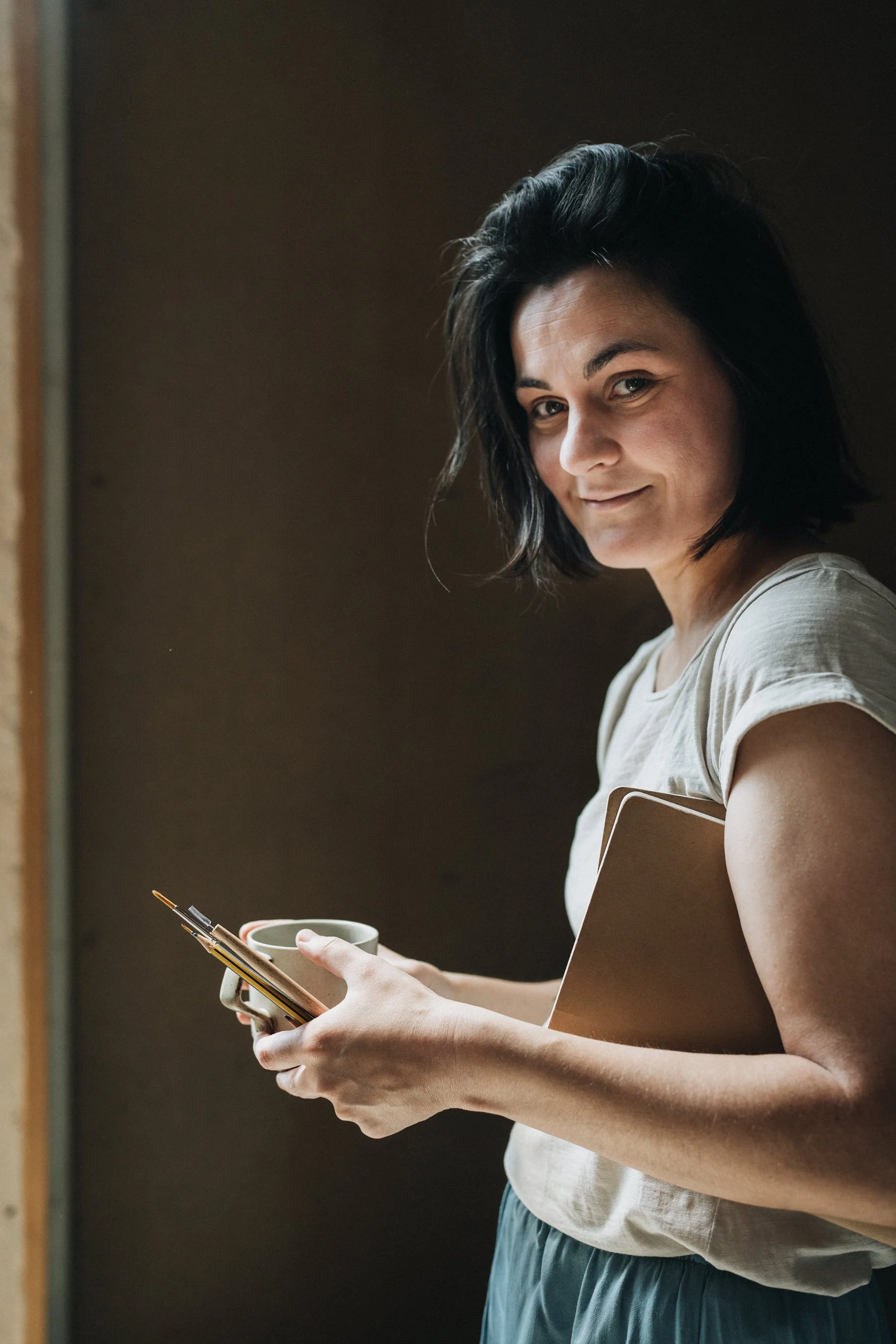 Jeune femme souriante avec des cheveux noirs, tenant une tasse, un carnet et un téléphone dans ses mains, debout devant un mur sombre.