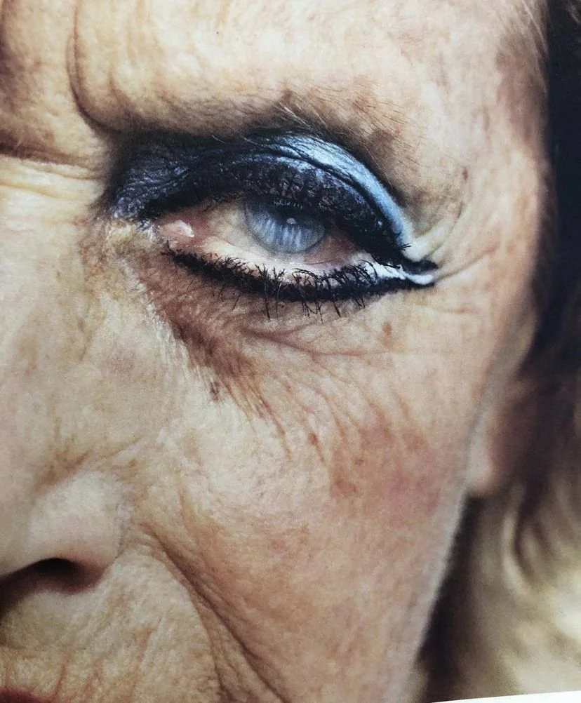 Un gros plan d'un œil de personne âgée avec maquillage noir et blanc autour de l'œil. La peau est ridée et fine.