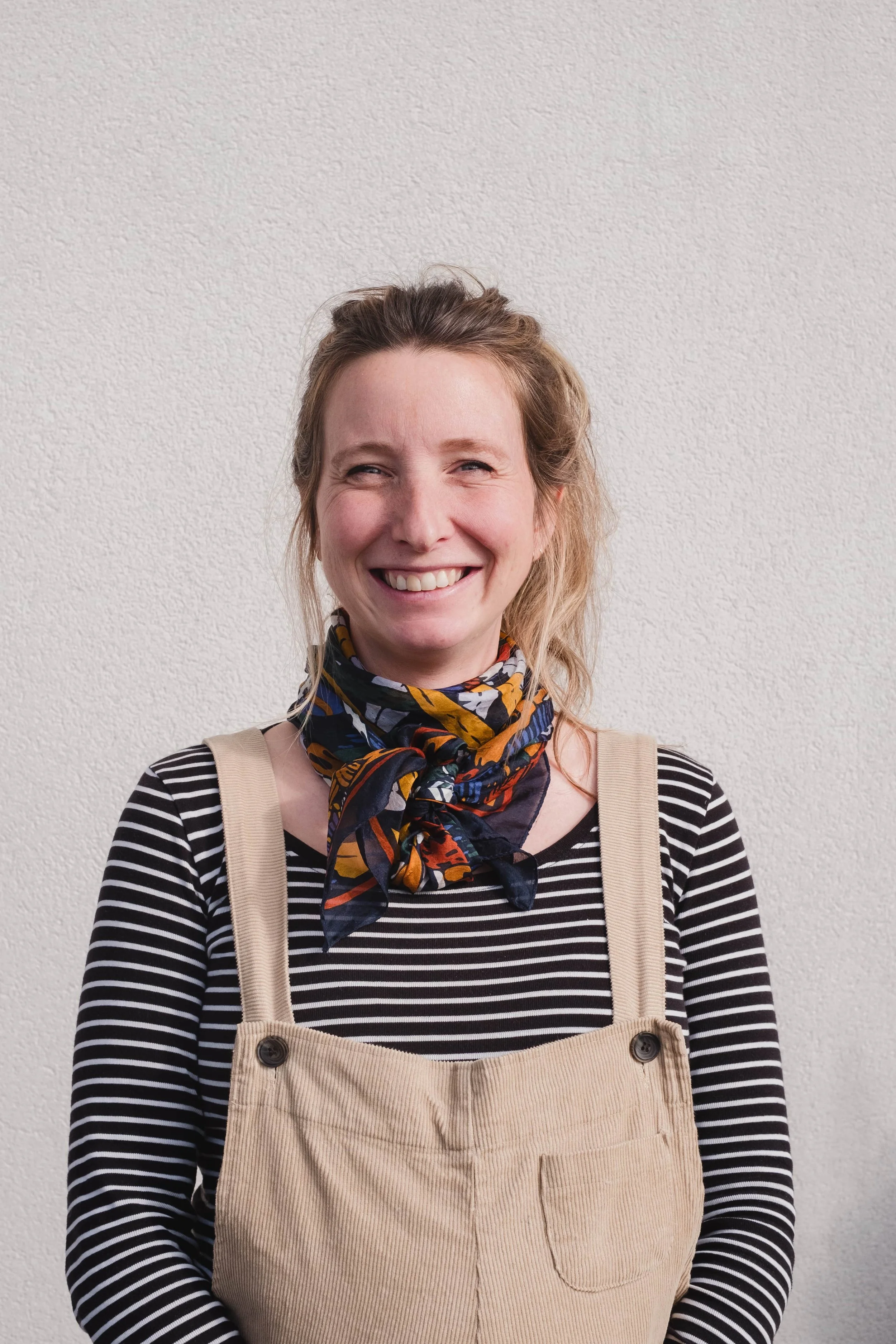Portrait d'une femme souriante portant un foulard coloré, un pull à rayures noires et blanches, et une salopette beige, debout contre un mur blanc.