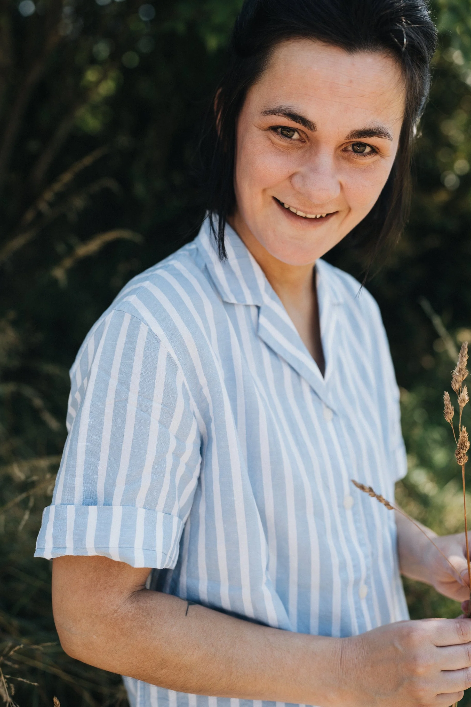 Une femme souriante, portant une chemise à rayures bleues et blanches, dans un environnement extérieur avec des plantes et un feuillage en arrière-plan.