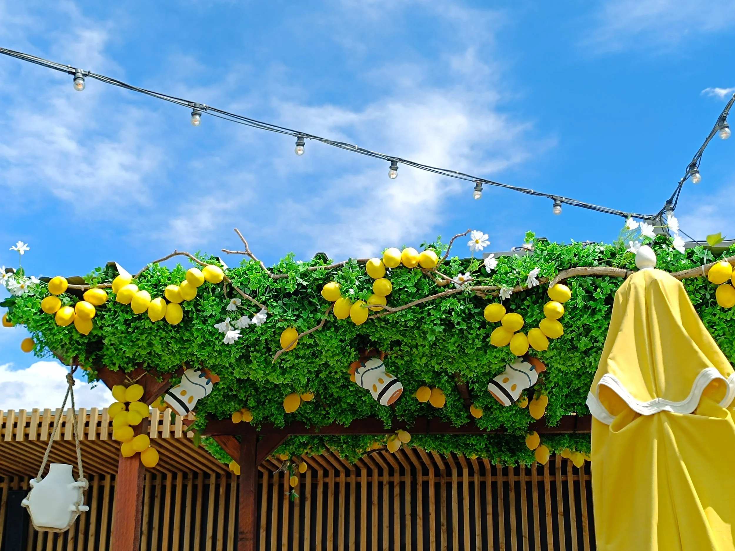 Décor de terrasse avec guirlandes lumineuses, fausse verdure, citrons jaunes, fleurs blanches, et éléments de décoration en céramique et tissu jaune contre un ciel bleu avec quelques nuages.