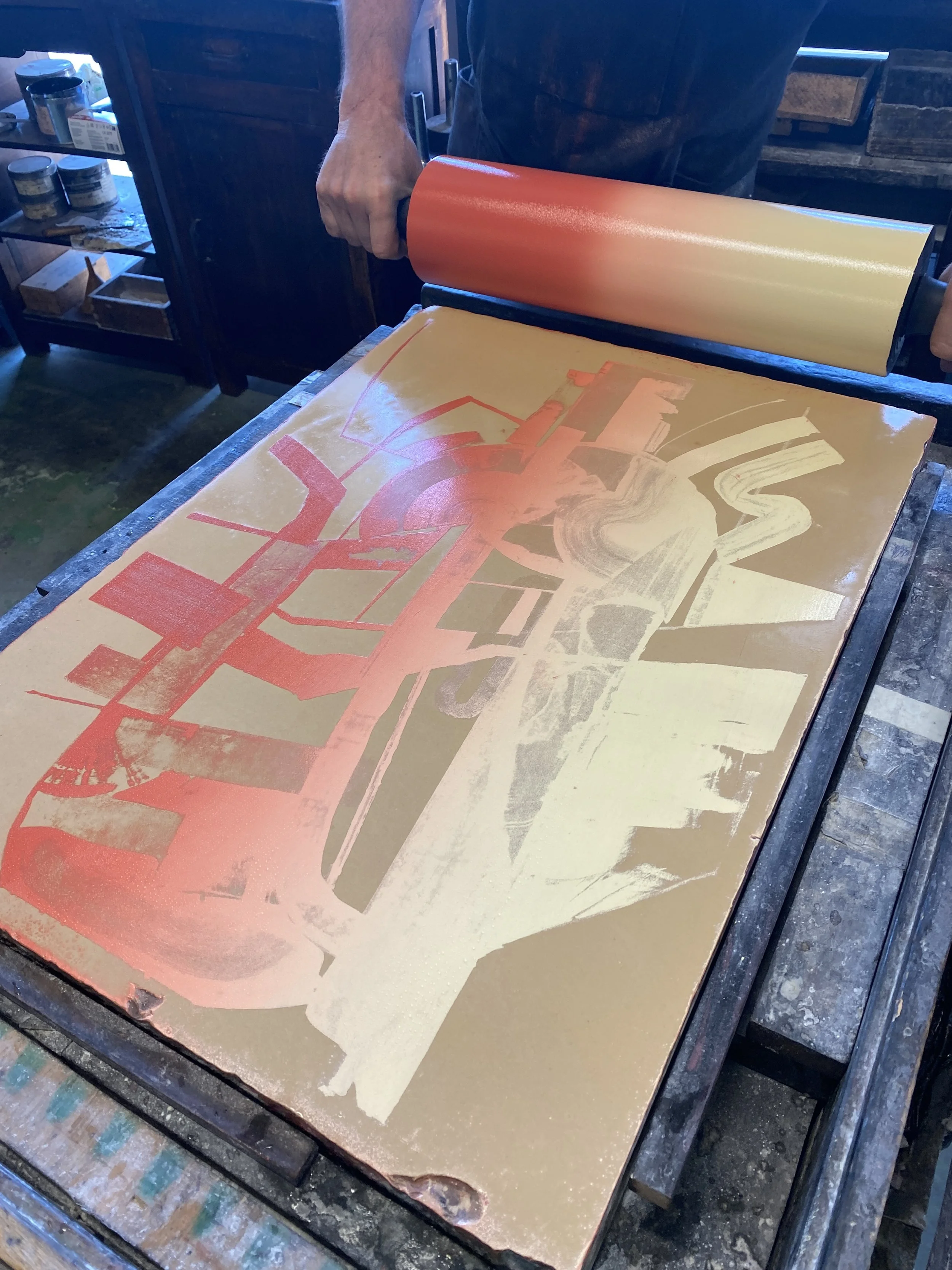 Une personne utilisant une presse pour créer une impression en lithographie avec des couleurs rouge, beige, jaune et blanche sur une surface plate.