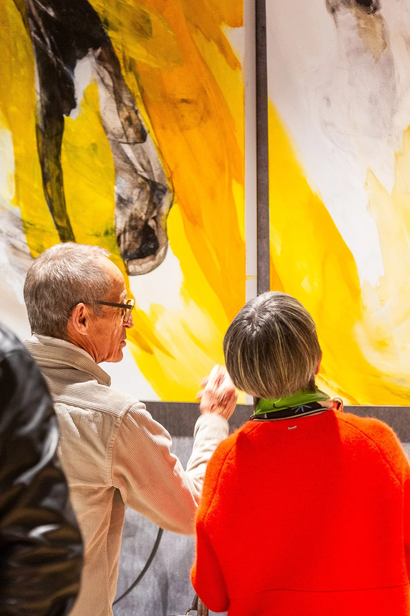 Deux personnes regardent une œuvre d'art abstraite dans une galerie, avec des couleurs jaune, orange, noir et blanc.