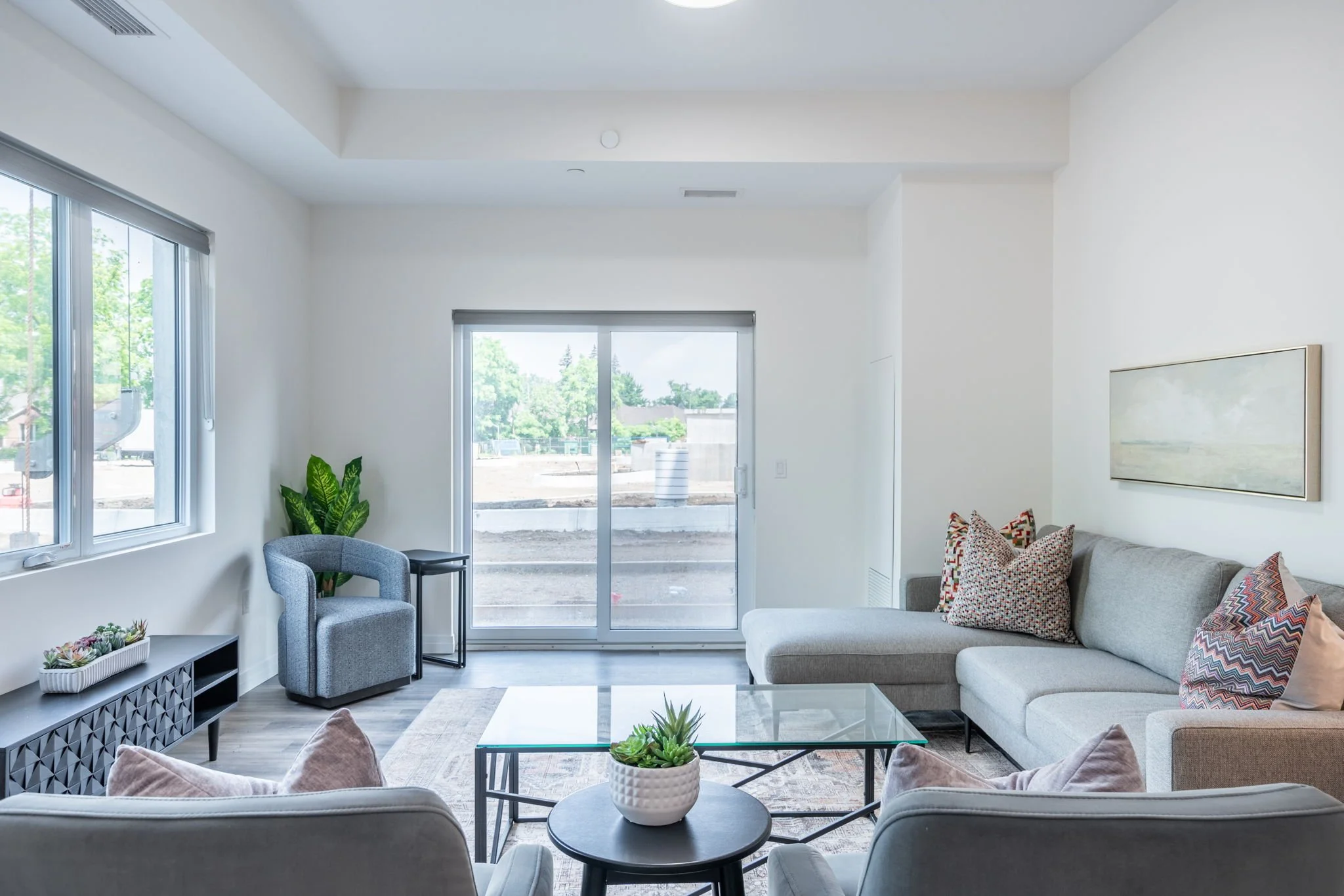 Living room  in St. Thomas, ON rental apartment with a gray sectional sofa, patterned pillows, a glass coffee table with a potted plant, a light-colored lounge chair, a small side table, large windows, a sliding glass door, and a landscape outside.