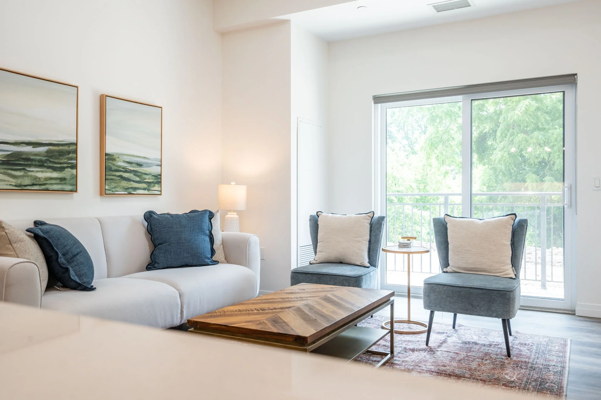 Living room in St. Thomas, ON rental apartment with a white sofa, blue pillows, a wooden coffee table, two blue armchairs with pillows, a floor lamp, sliding glass door with a balcony, and wall art with green landscape paintings.