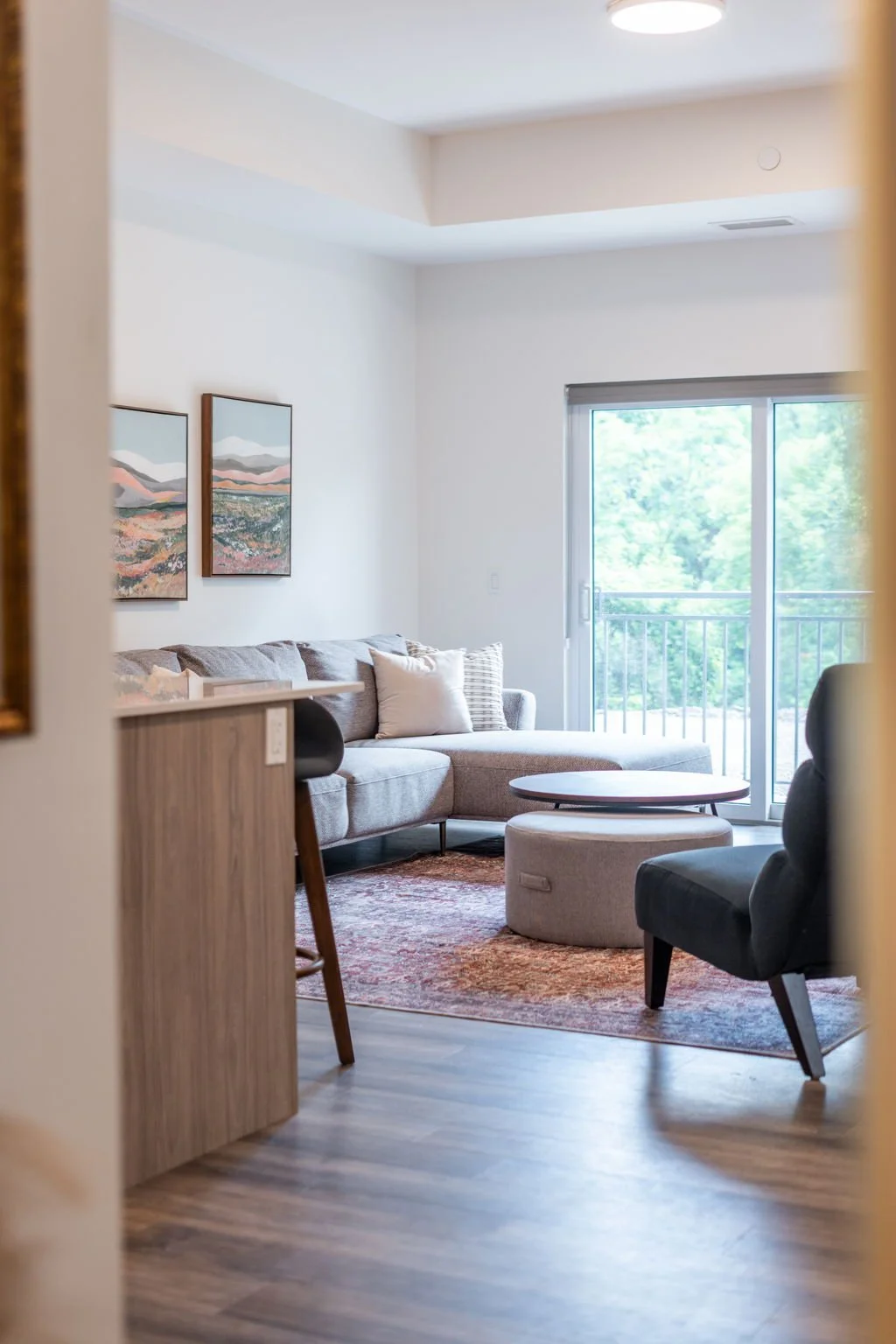 Living room in St. Thomas, ON rental apartment with gray sectional sofa, black and beige chairs, round coffee table, sliding glass door with balcony, and abstract mountain landscape paintings on white wall.