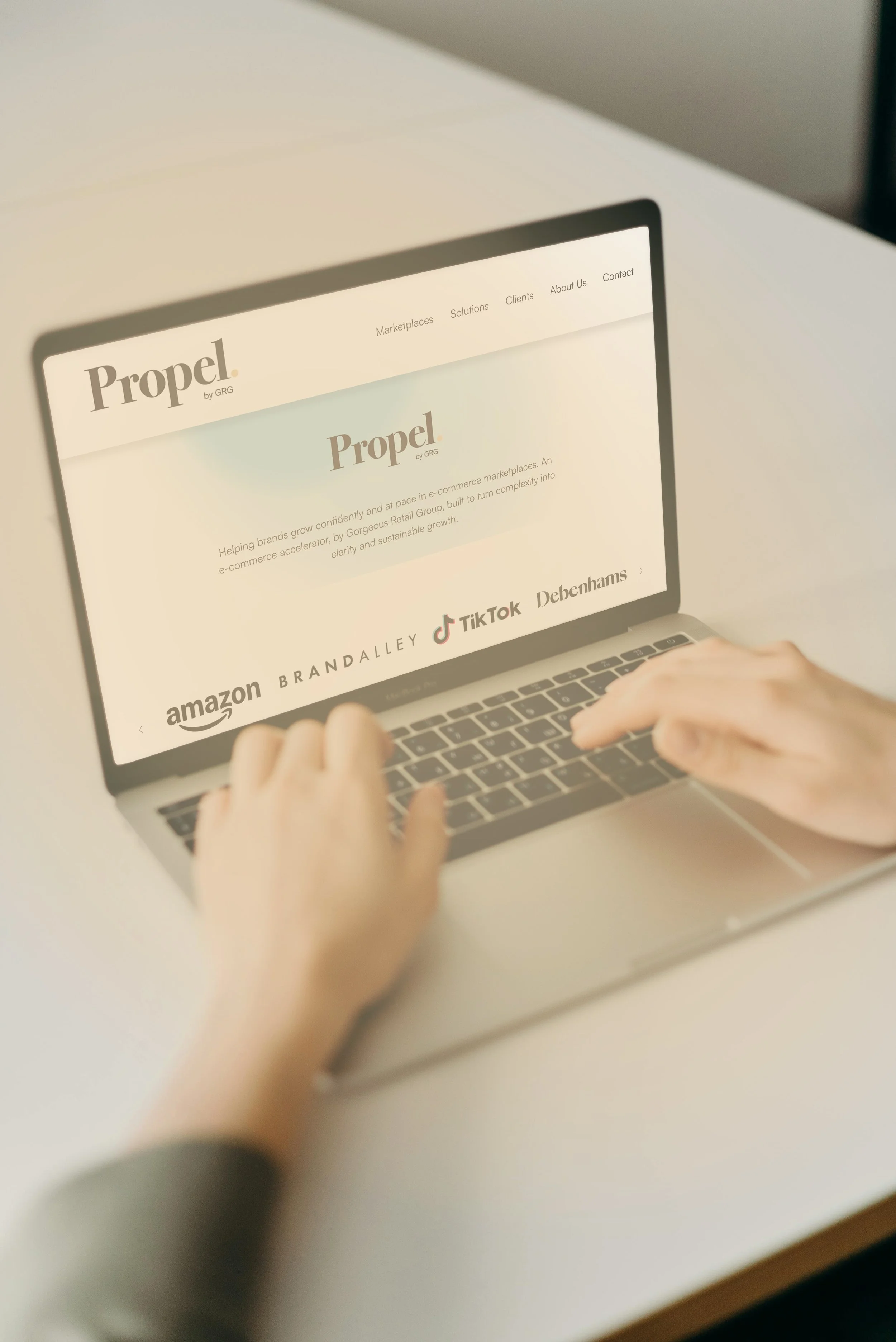 A person using a laptop on a white desk, browsing a website with the logo 'Propel by GRG' and various navigation links at the top.