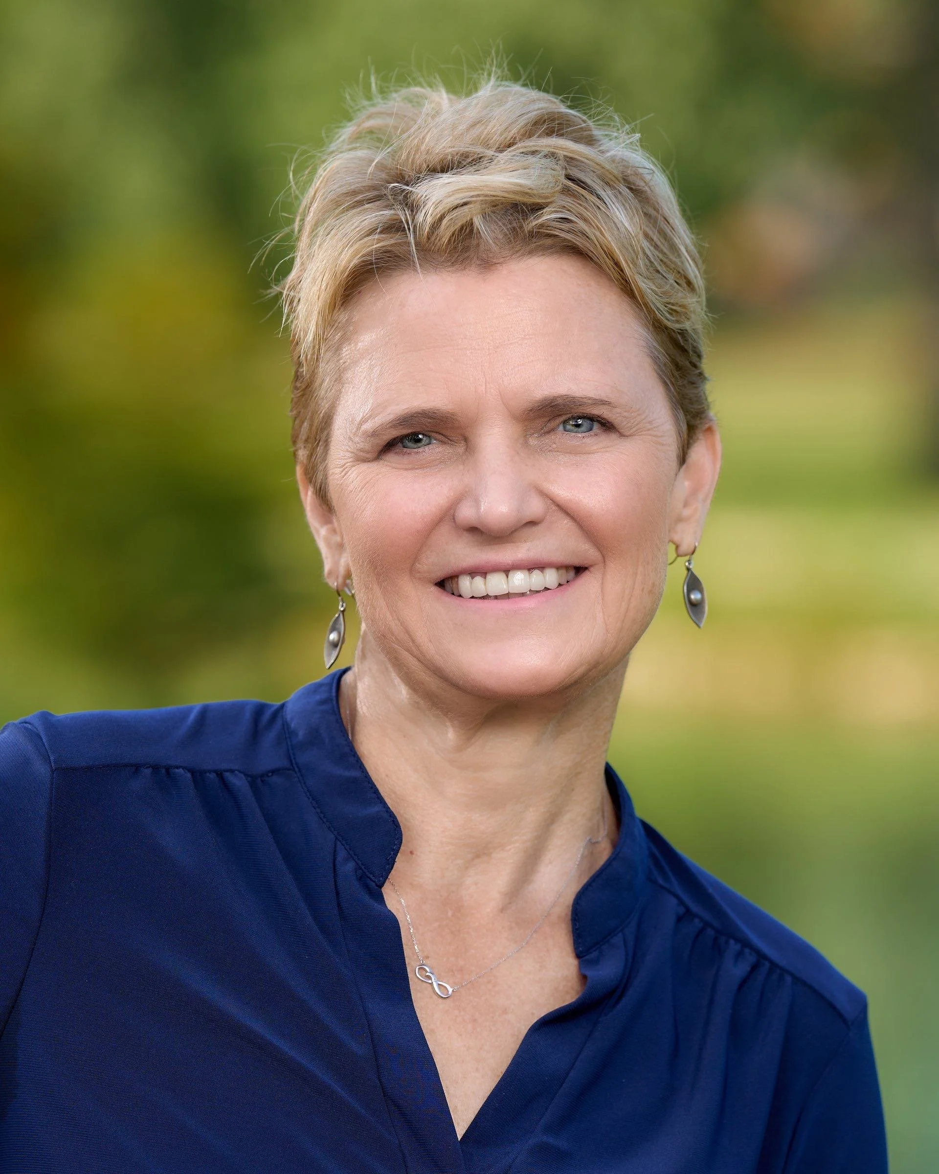 Close-up photograph of a smiling middle-aged woman with short blonde hair, wearing a blue blouse, earrings, and a necklace, outdoors with a blurred green background.