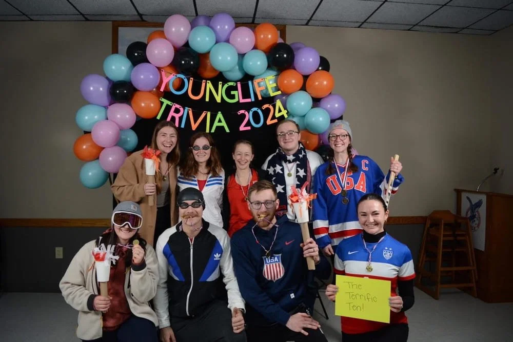 Group of adults smiling in front of a colorful balloon arch with a sign that reads "Younge Life Trivia 2024." 