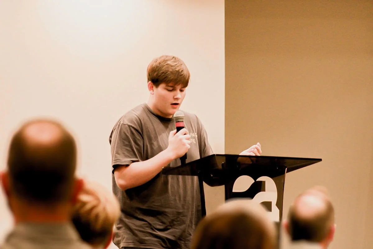 A young boy standing at a podium, speaking about the positive impact volunteering adults made in his life through teen fellowships and events.
