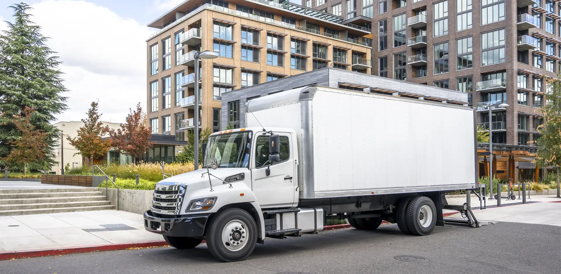 White box truck parked on a city street in front of modern apartment buildings and landscaped area with trees.