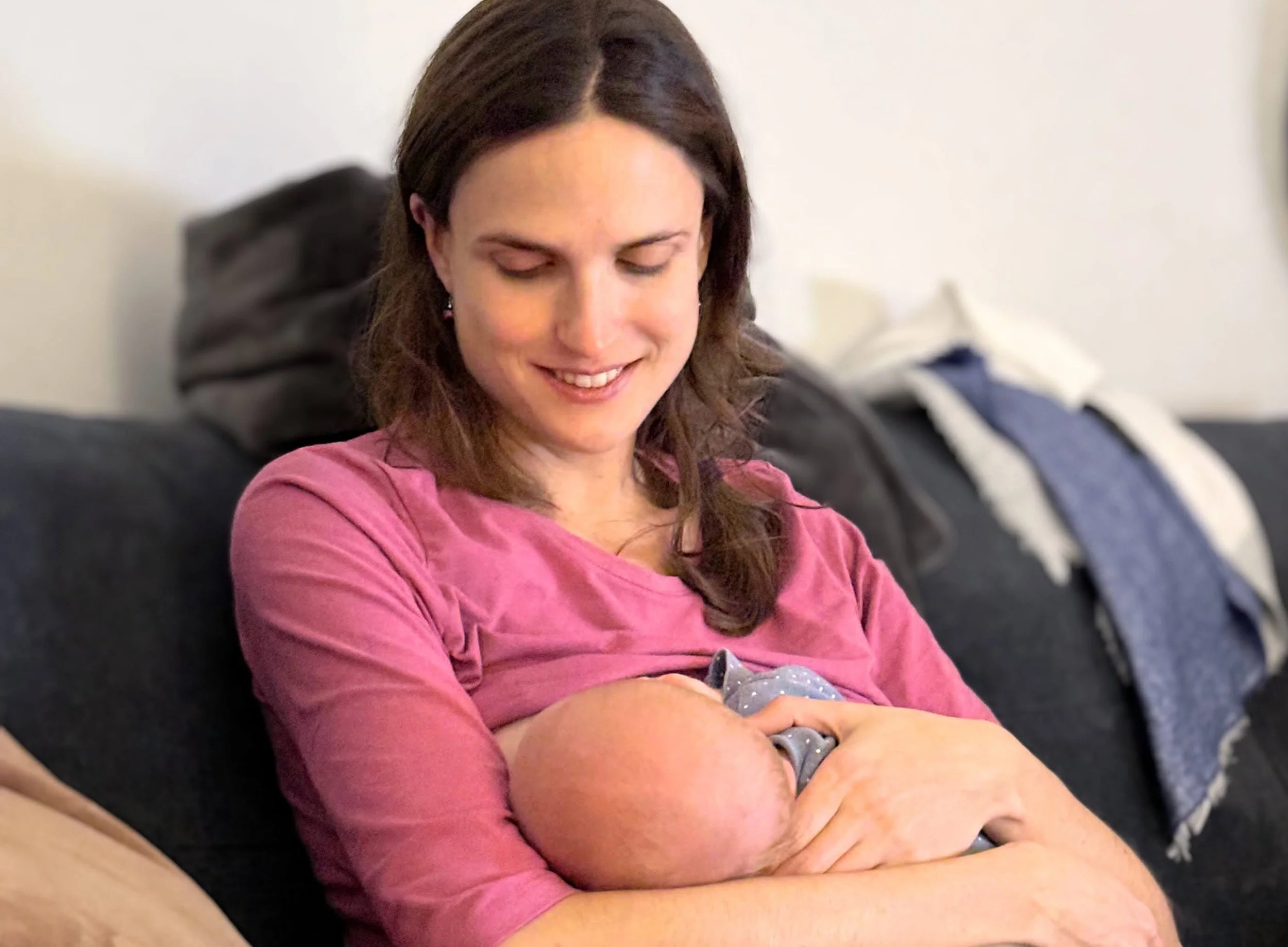 A woman with brown hair wearing a pink shirt is smiling and looking down at a sleeping baby she is holding in her arms.