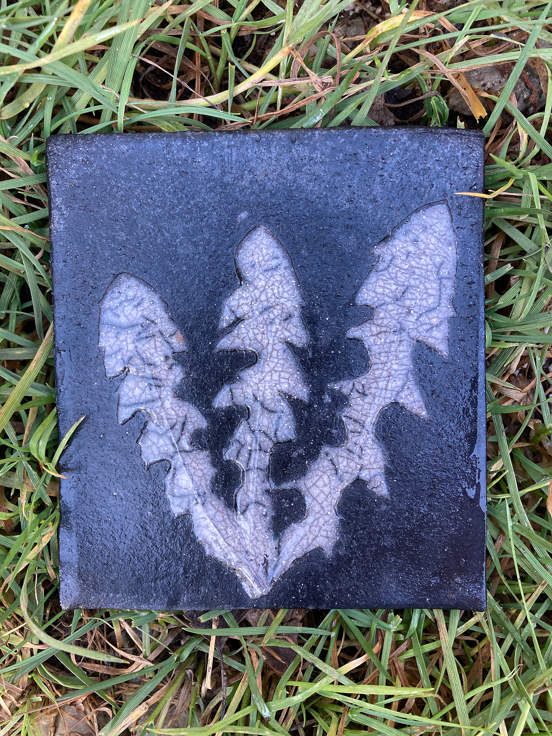 Eine quadratische, Keramik-Kachel mit Raku mit einem weißen, detaillierten Abbild von einem Farnblatt, liegt auf Gras. Die Platte hat eine raue Oberfläche.