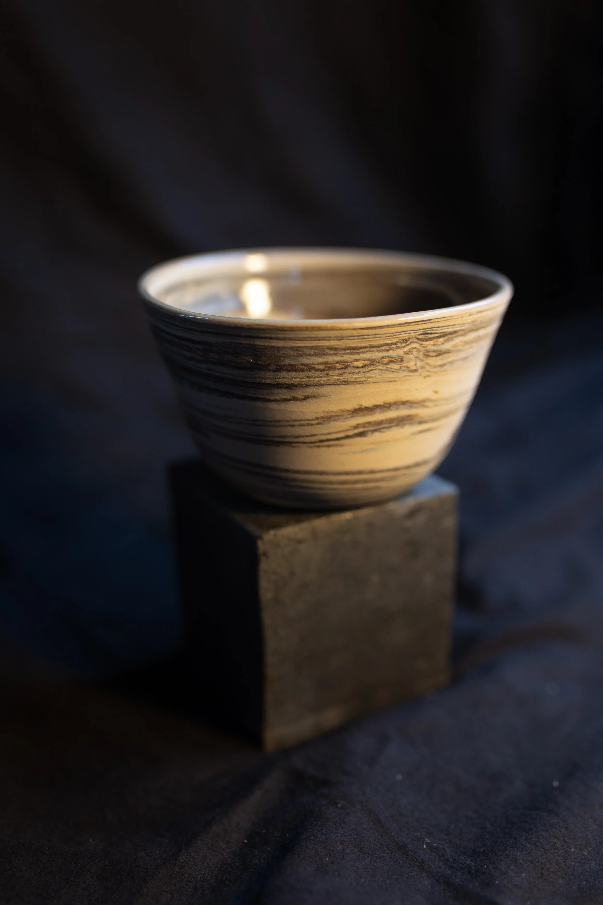 Kleine keramische Schale mit schwarzen und beigen Streifen auf einem schwarzen Würfel, dunkler Hintergrund.