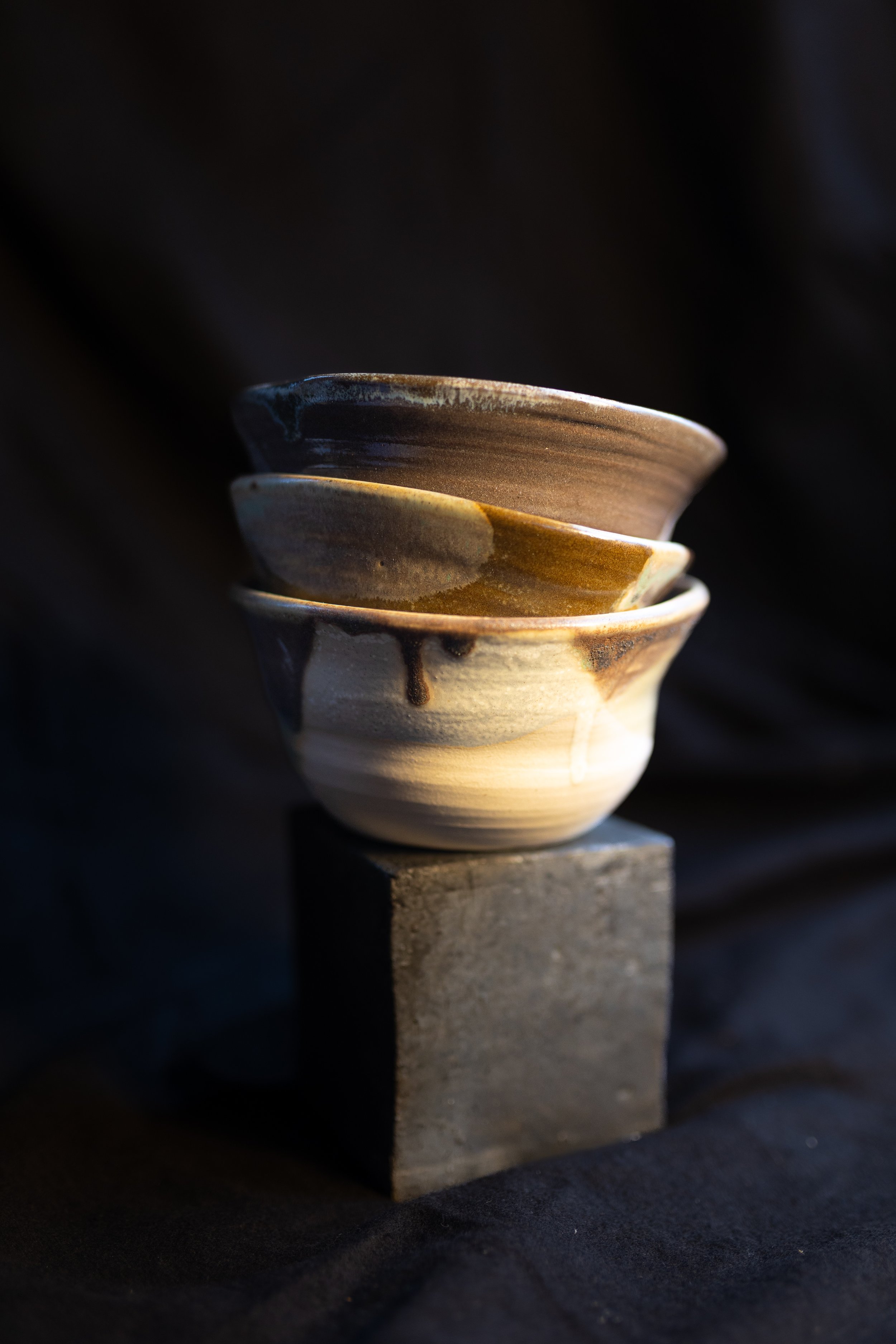 Drei handgefertigte Keramikschalen in Erdtönen, gestapelt auf einem schwarzen Block, auf dunklem Hintergrund.