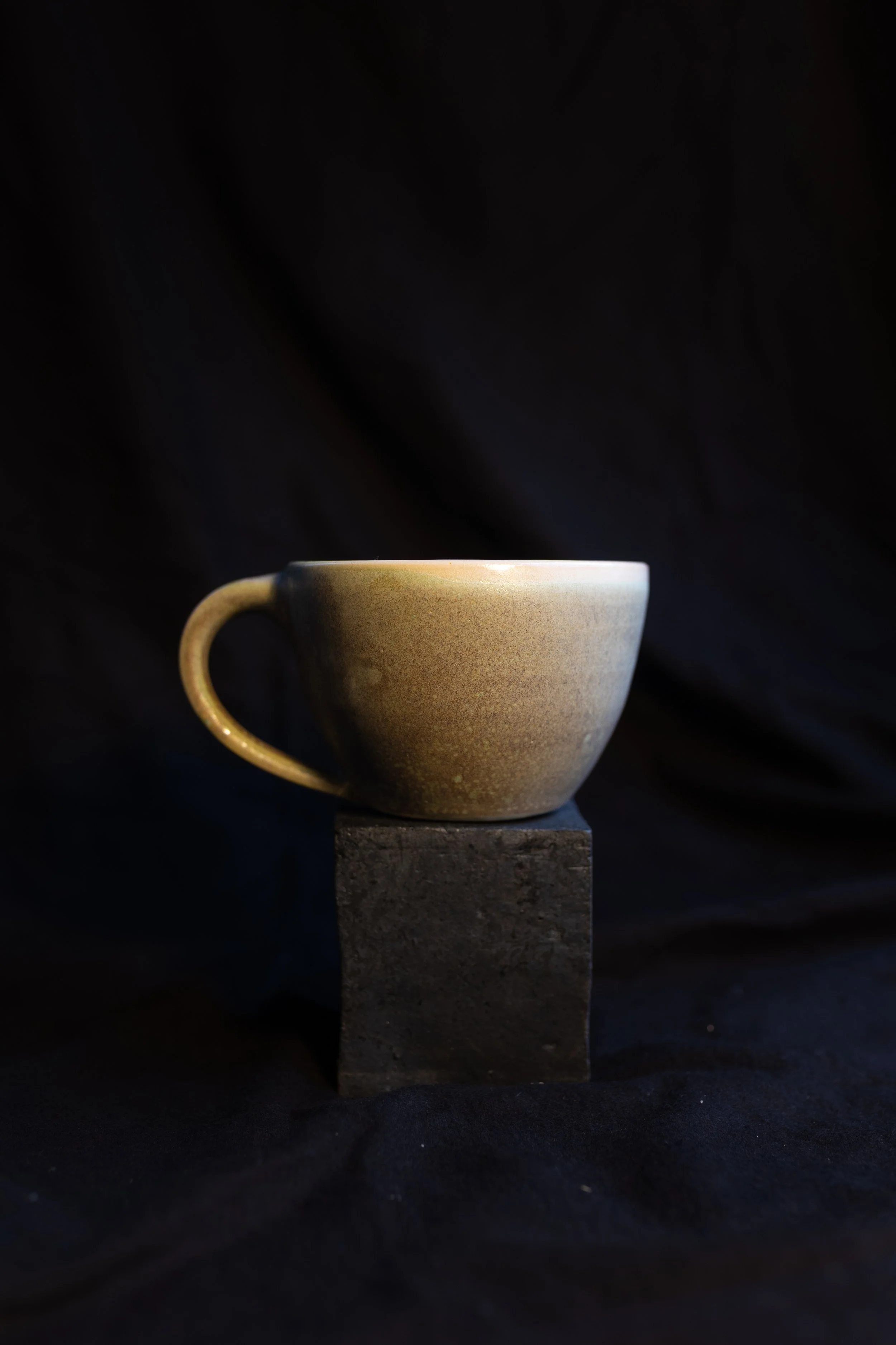 Eine Kaffeetasse auf einem schwarzen Block, vor schwarzem Hintergrund.