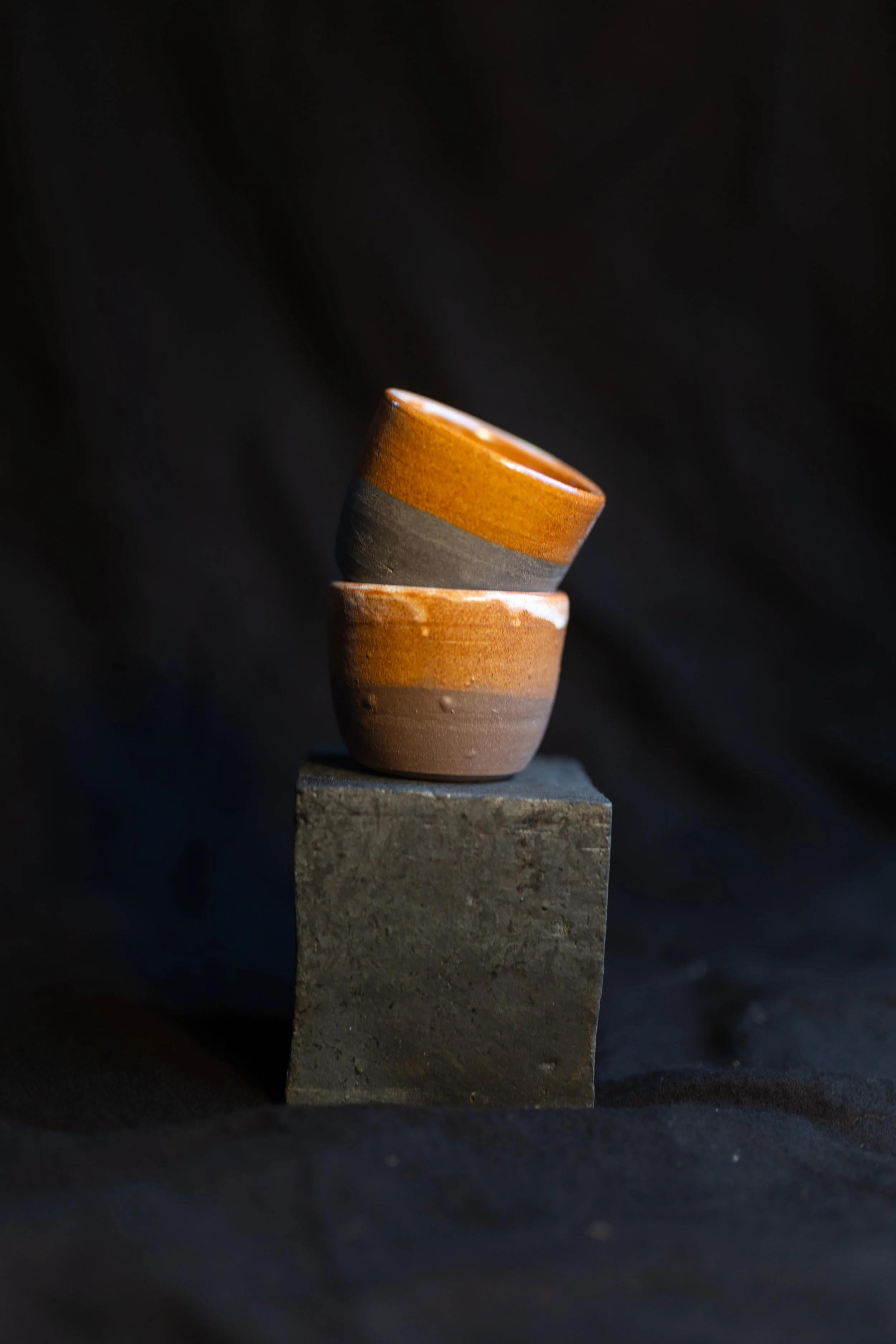 Zwei handgefertigte Keramiktassen, die übereinander auf einem schwarzen Steinquader stehen, vor einem schwarzen Hintergrund.