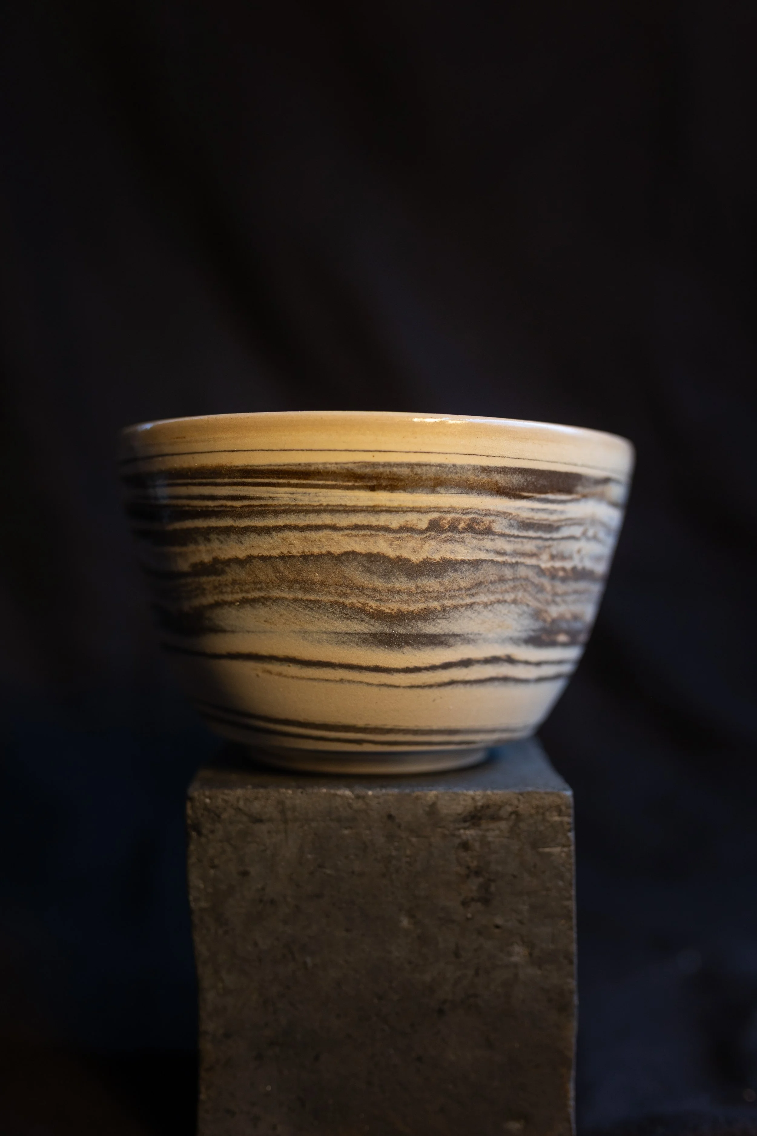 Keramikschale mit braunen und beigen Streifen auf einem Steinblock gegen schwarzen Hintergrund.