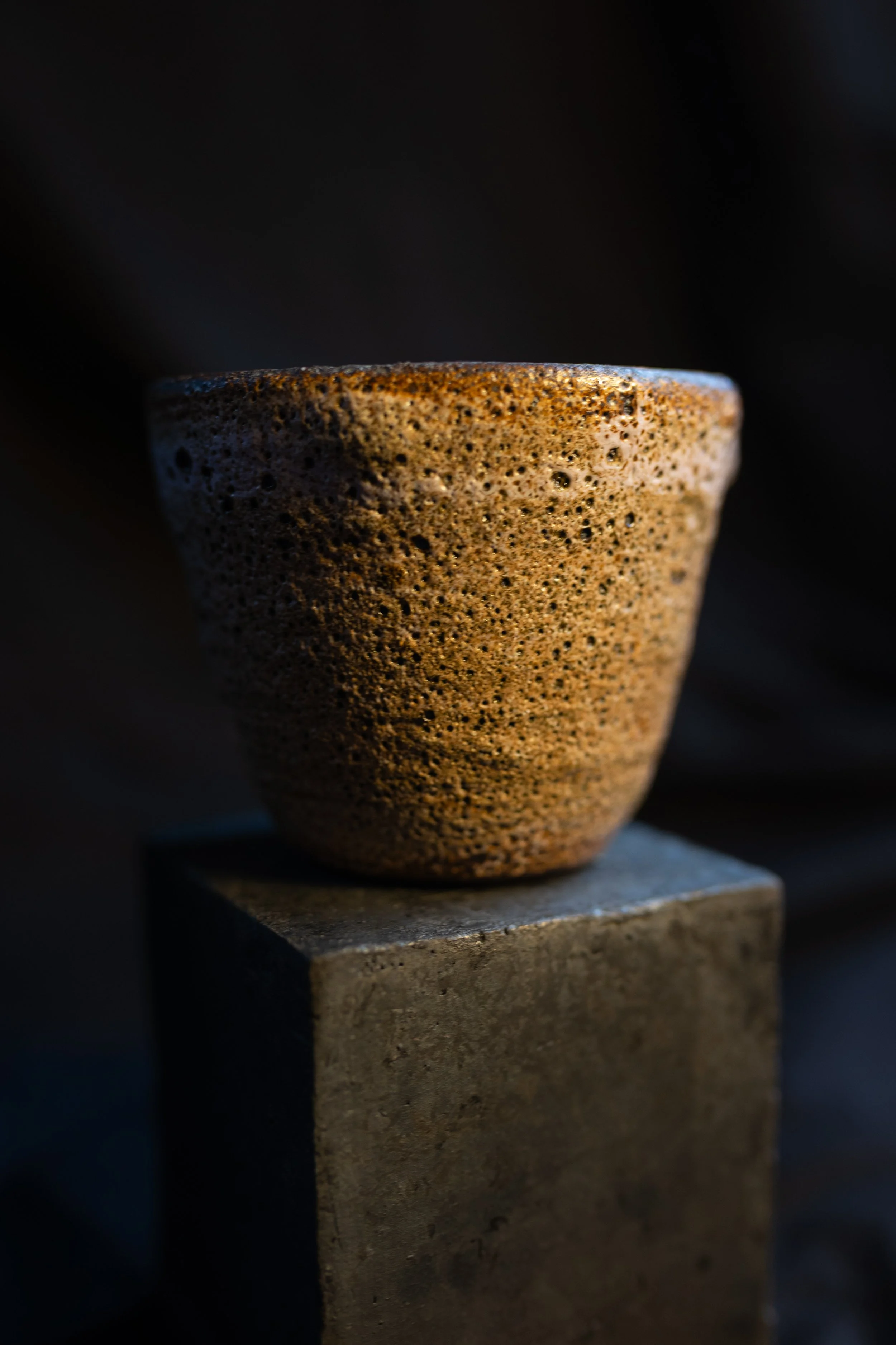 Eine Tasse mit einer rauen, porösen Oberfläche auf einem quadratischen Sockel, dunkler Hintergrund.