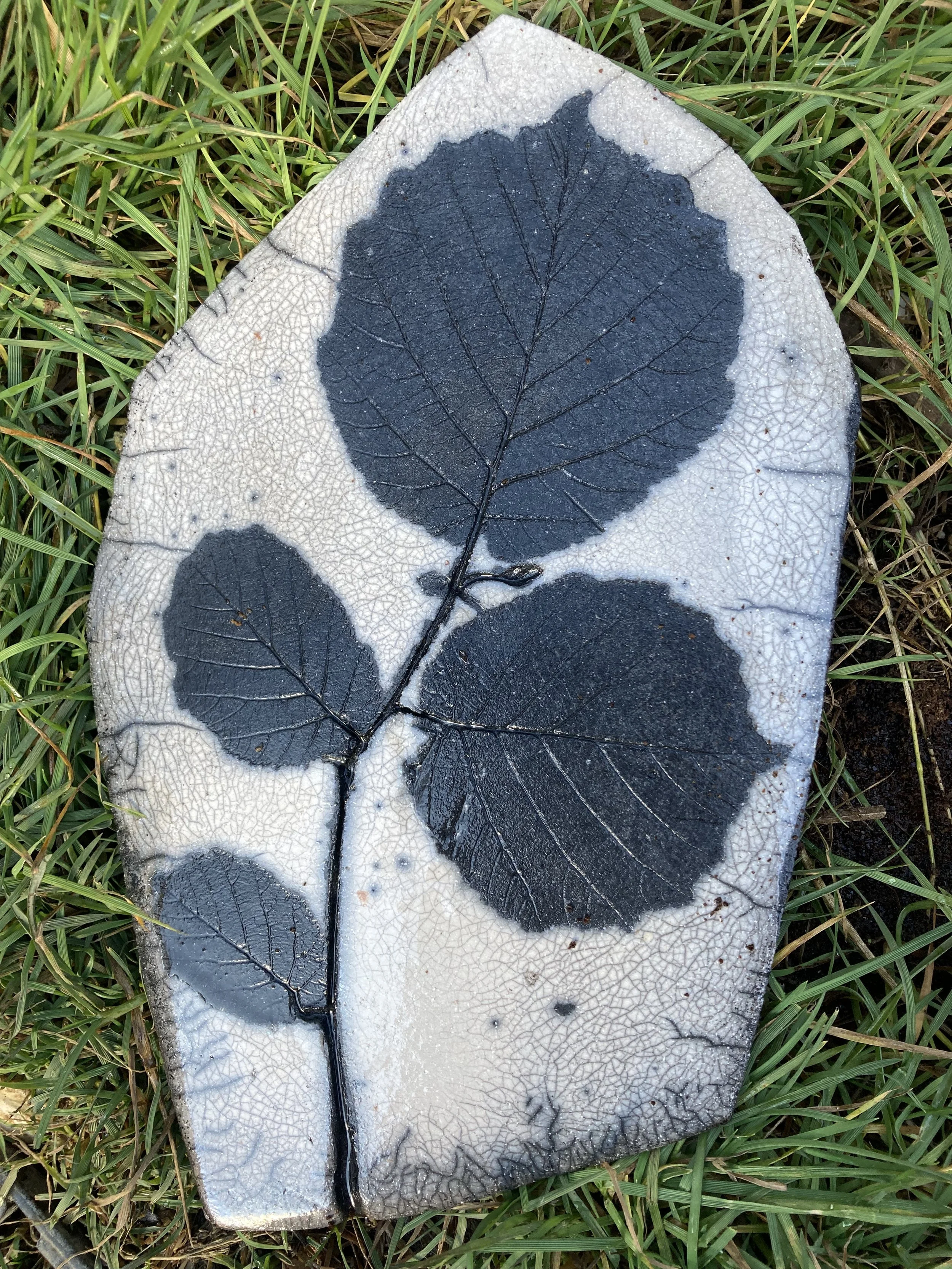 Eine Keramik-Kachel mit Raku von schwarzen Blätternmuster auf weißem Hintergrund, liegend im Gras.