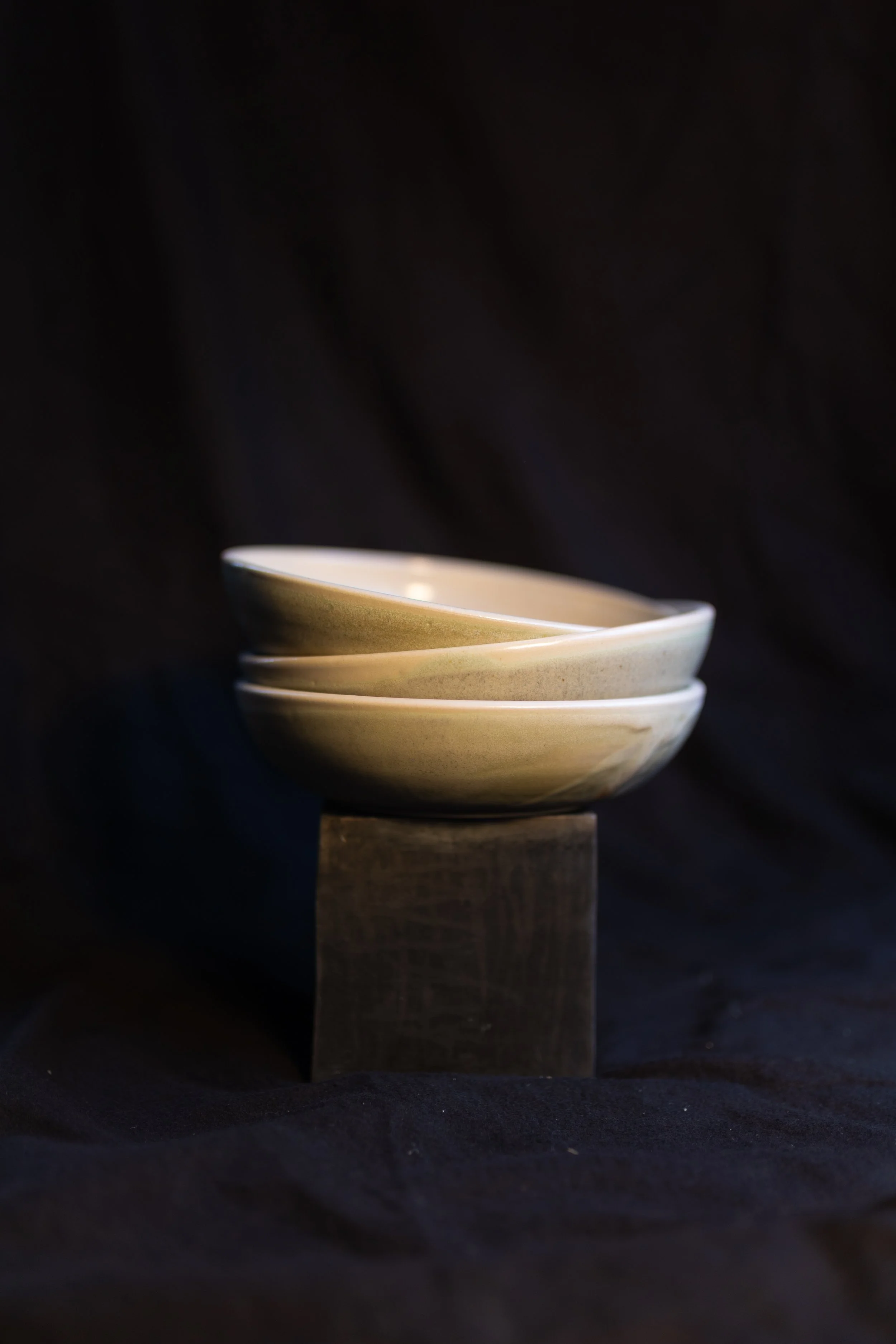 Drei beige Keramikschalen, gestapelt auf einem dunklen Holzblock vor einem schwarzen Hintergrund.