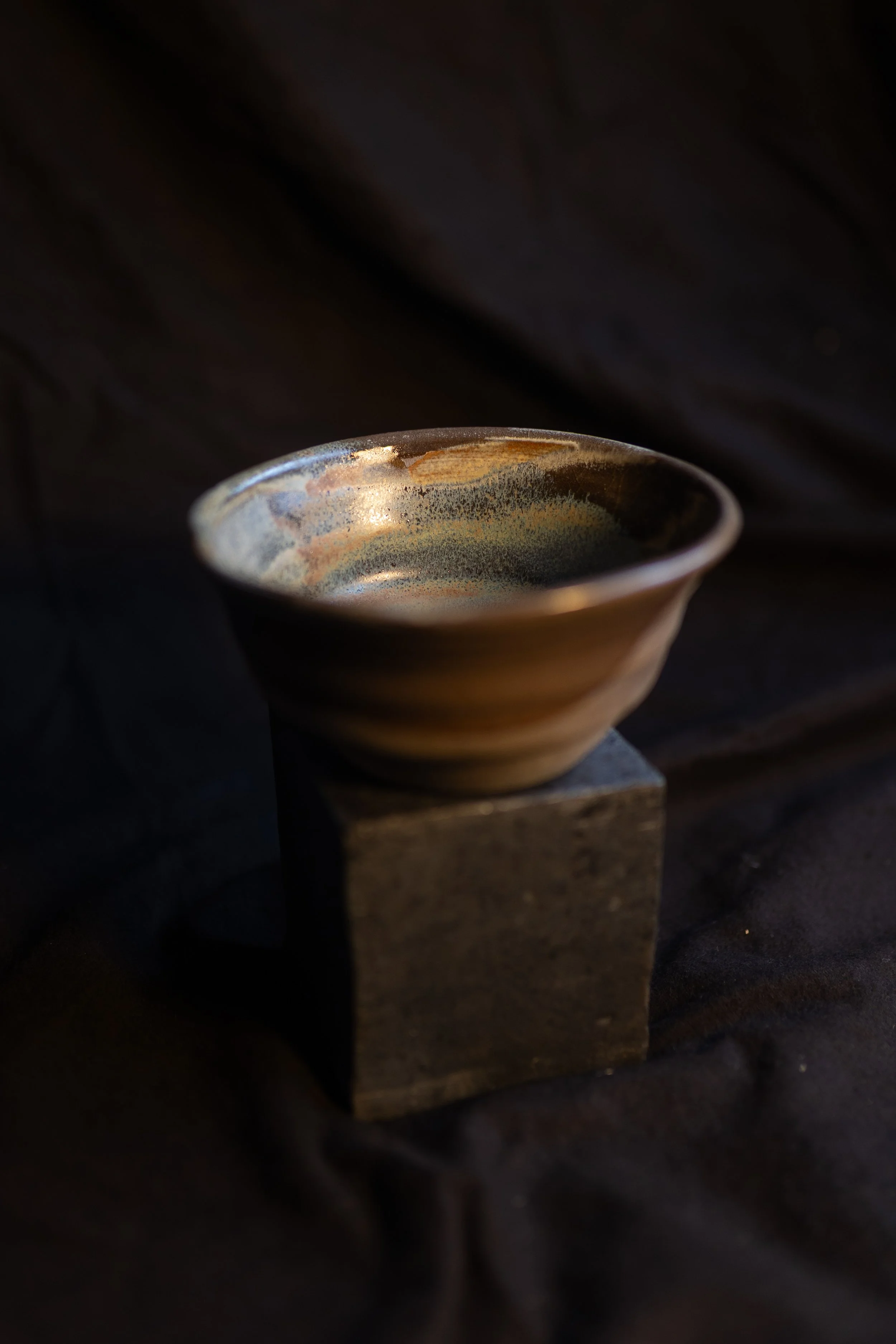 Eine Keramikschale mit dunklen, warmen Farben auf einem schwarzen Holzblock vor schwarzem Stoffhintergrund.