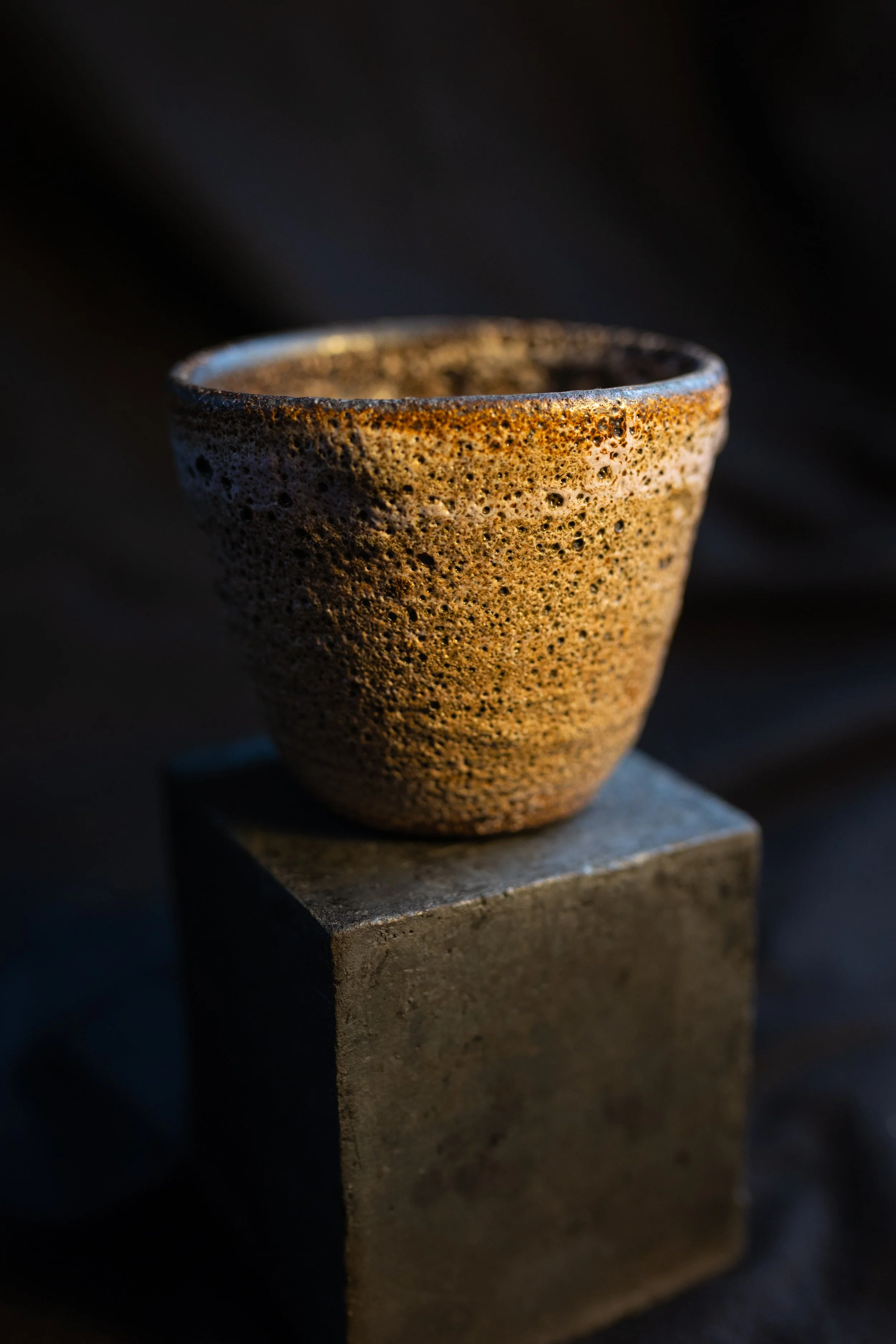 Ein grober, keramischer Becher mit brauner, sandartiger Oberfläche auf einem grauen Sockel, dunkler Hintergrund.