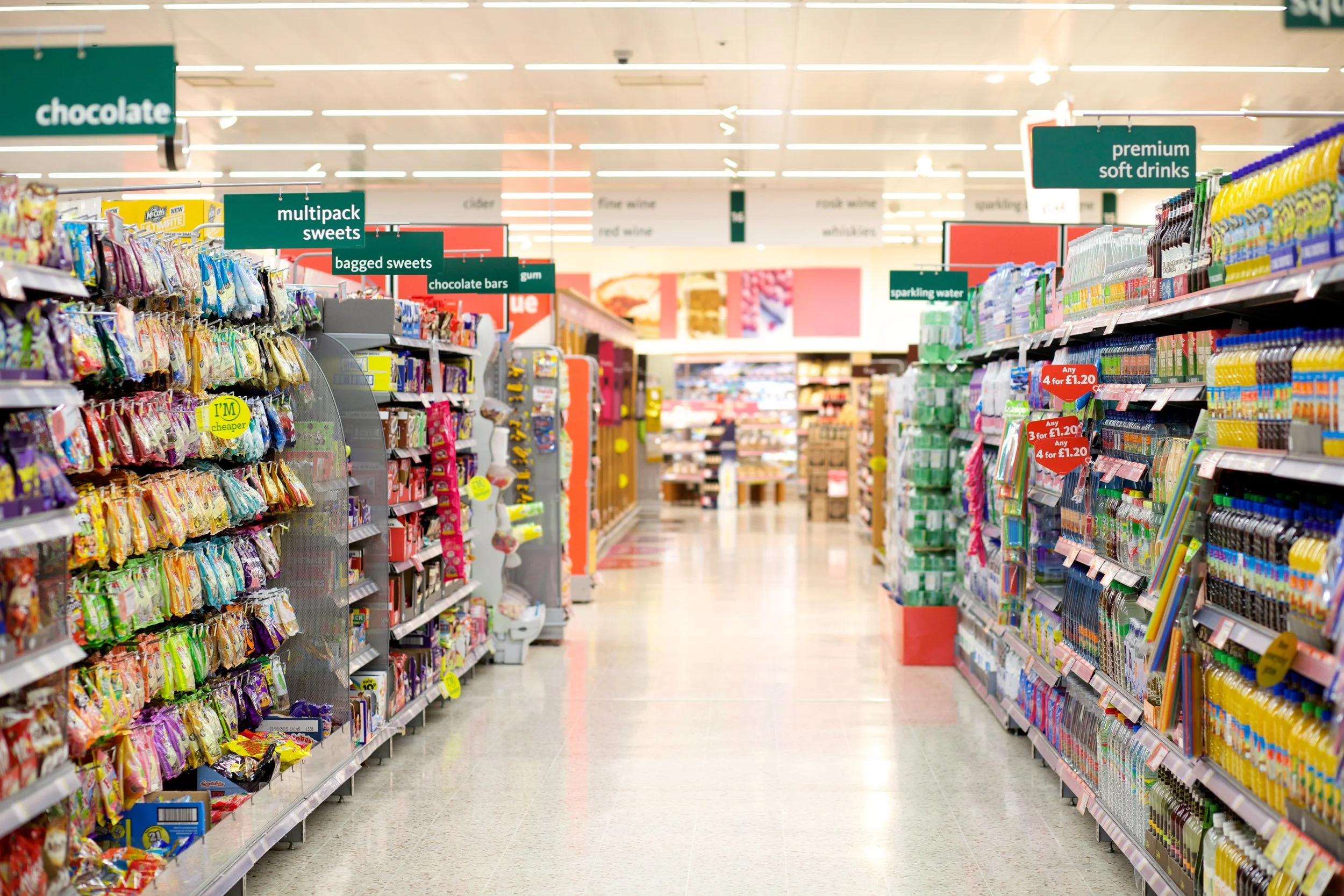 A grocery store aisle with shelves stocked with various snacks on the left and beverages on the right. Signs hang from the ceiling indicating sections such as 'multipack sweets,' 'bagged sweets,' 'chocolate bars,' 'premium soft drinks,' and 'sparkling water.' The floor is shiny and reflective, extending toward a back section with more shelves and products.