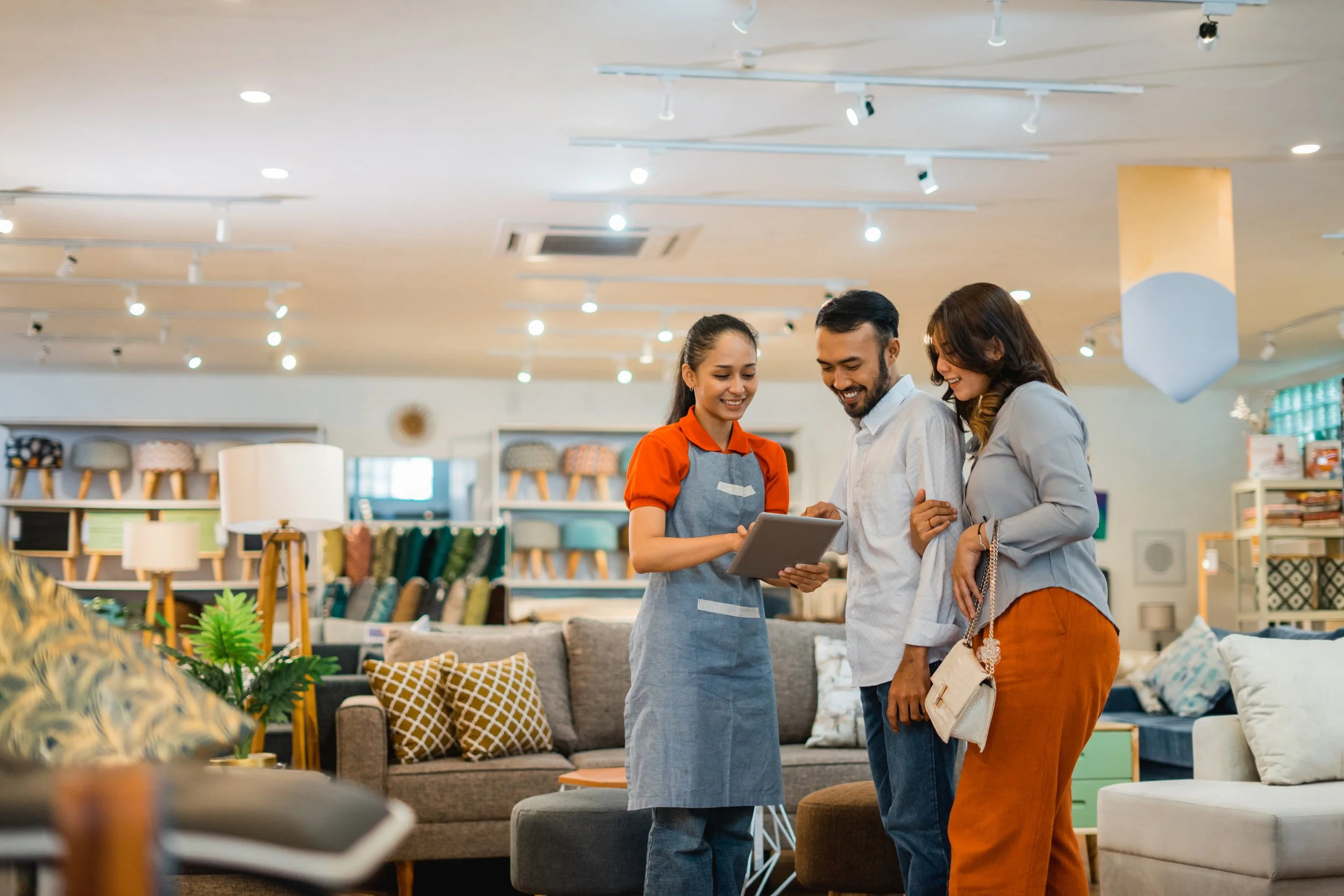 A sales associate showing a tablet to a smiling couple inside a furniture store with sofas and lighting fixtures in the background