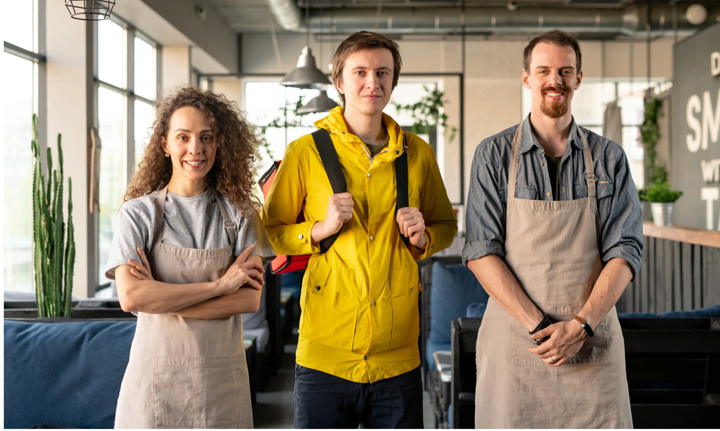 Three people standing in a cafe or restaurant, two women wearing aprons and a man wearing a backpack and yellow raincoat, all smiling at the camera.
