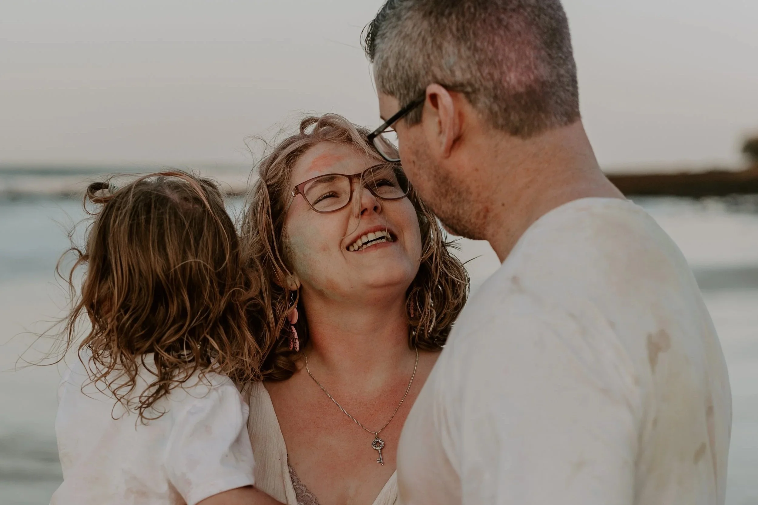 Smiling woman with glasses holding two children on a beach, a man facing her close up.